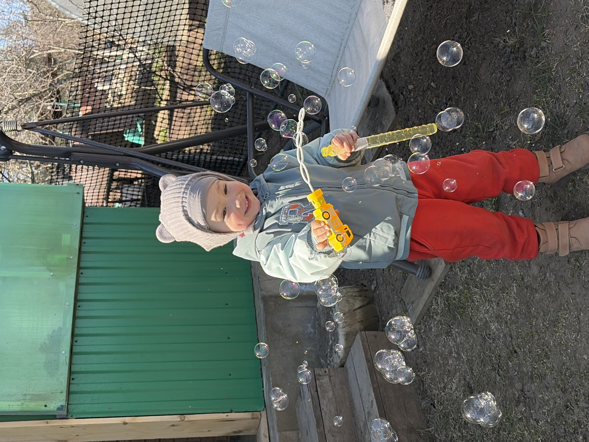 The image captures a child playing with bubbles outdoors, likely in a backyard setting. The child, wearing a grey knitted hat, a light blue jacket, red pants, and tan boots, is holding two bubble-making devices: a yellow car-shaped bubble blower and a yellow wand. Numerous iridescent bubbles float around the child, suggesting active bubble blowing.
The background consists of several elements. A green corrugated metal fence is visible to the left. A light grey outdoor chair is situated behind the child. The ground appears to be covered in grass and some bare soil. A metal swing is seen in the background behind the fence.
The specific location of Chaikovsky, Russia, cannot be verified from the visual information provided.