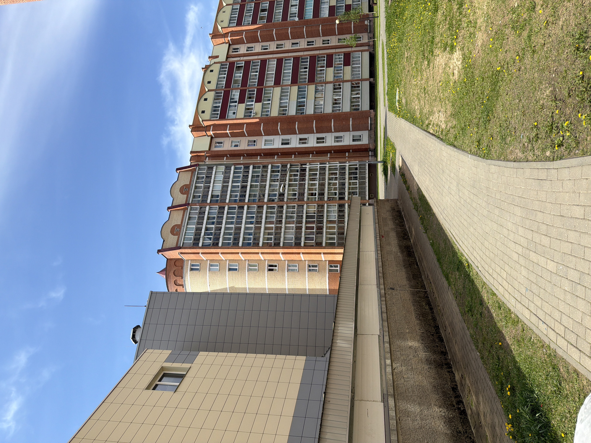 The image shows a view of buildings and a walkway in an urban area of Unknown, Unknown. On the left, there is a low-rise building with tiled beige and grey exterior walls and at least one visible window. Behind it is a tall apartment building with a mix of brick, painted, and windowed sections. The walkway is paved with grey bricks and runs parallel to a narrow strip of grass with yellow flowers. The sky is blue with light clouds.
