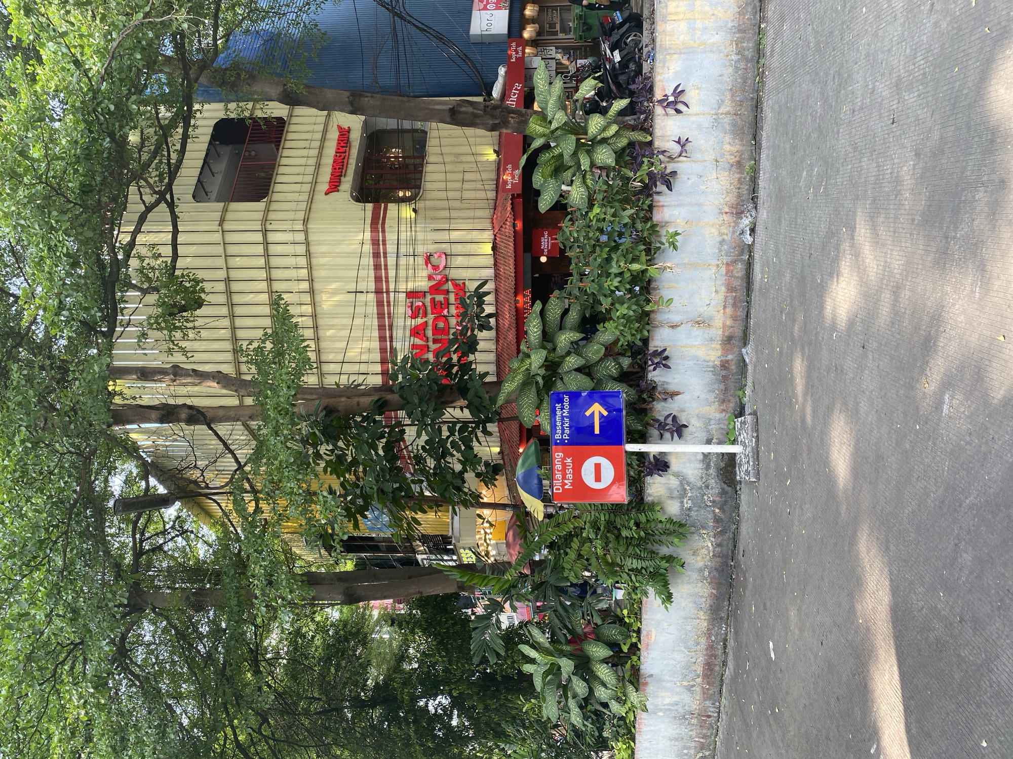 The image depicts the exterior of a building constructed from shipping containers in the bustling city of South Jakarta, Indonesia. The building, painted in shades of yellow and red, appears to be a commercial establishment, possibly a restaurant or cafe, as indicated by the prominent signage reading "NASI RENDANG" and "MINANG PRIDE." Lush green foliage, including large-leafed plants and trees, surrounds the base of the building, creating a vibrant contrast with the urban environment. In the foreground, a concrete road leads towards the building, with a directional sign indicating "DILARANG MASUK" (Do Not Enter) and an arrow pointing towards "Basement - Parkir Motor" (Basement - Motorcycle Parking). The overall scene suggests a lively urban setting, characteristic of South Jakarta, with a blend of modern architecture and natural elements.