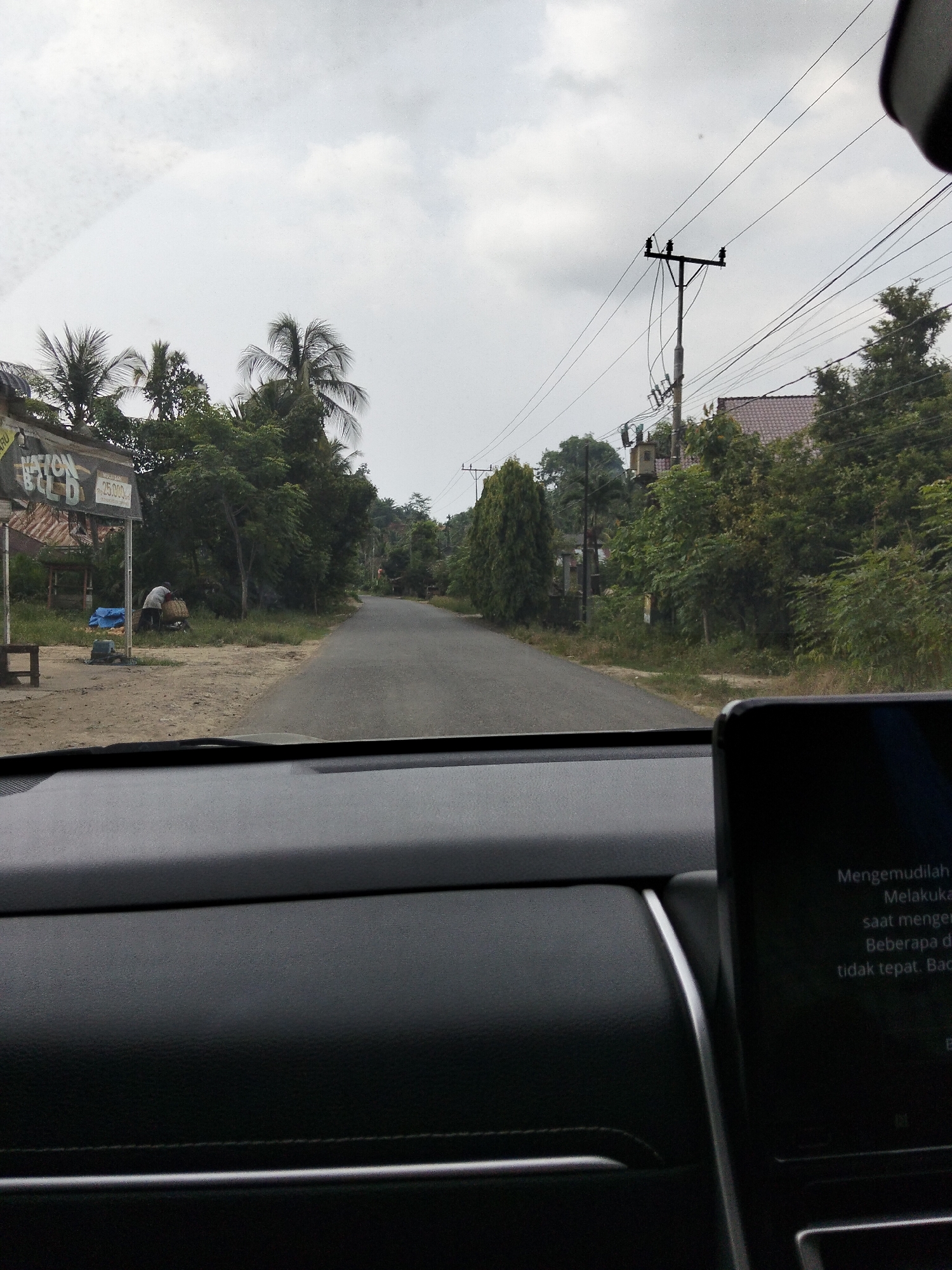 The image captures a view from inside a vehicle, looking out onto a two-lane paved road in Blang Mee, Indonesia. The sky is overcast, with visible clouds. On the left side of the road, there's a roadside stall or building with a sign that is partially visible and reads "NATION BLD." Adjacent to this structure, a person is observed standing next to a large basket, seemingly engaged in some activity. Several blue tarps are laid out on the ground. Various trees, including palm trees, line the road. Further down the road, a utility pole with wires and a transformer is visible. On the right side of the road, houses and more trees can be seen. The interior of the vehicle shows a dashboard with a digital display screen, which has text in what appears to be Indonesian.