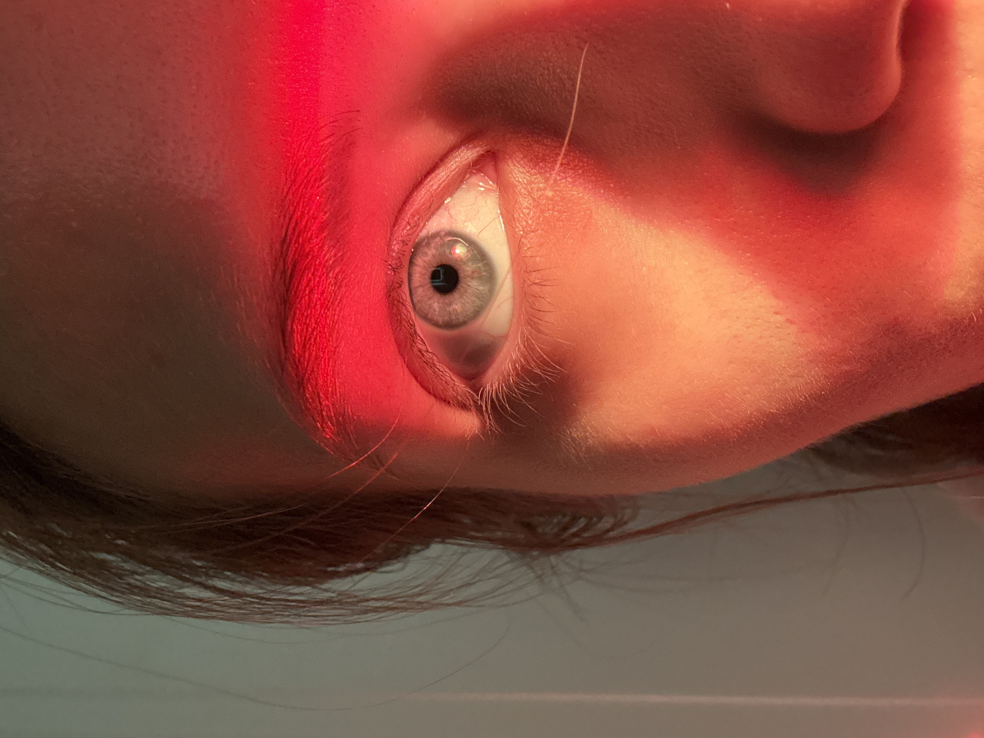 A close-up, rotated shot focuses on a person's left eye (from the viewer's perspective), illuminated by a strong red light beam. The bright red light intensely highlights the skin around the eye and the eyebrow, creating a dramatic contrast with the shadows on the cheek and forehead. The person's skin shows visible texture, including pores and fine hairs, accentuated by the harsh lighting.
The eye itself has a light-colored iris, appearing grey or light blue-green, with a prominent dark pupil. A small, bright, square-like reflection is visible within the pupil, suggesting an artificial light source. The sclera (white of the eye) shows faint red blood vessels. Eyelashes are visible, especially on the lower lid, and part of the eyebrow is strongly lit by the red beam.
The overall composition gives an impression of an examination or a deliberate artistic setup due to the focused and intense lighting. The person's face is cropped tightly, with hair visible along the bottom edge, suggesting a head tilted downwards or the camera angled upwards. No specific activity beyond the eye being open under the light can be clearly identified. There is no visible text in the image.