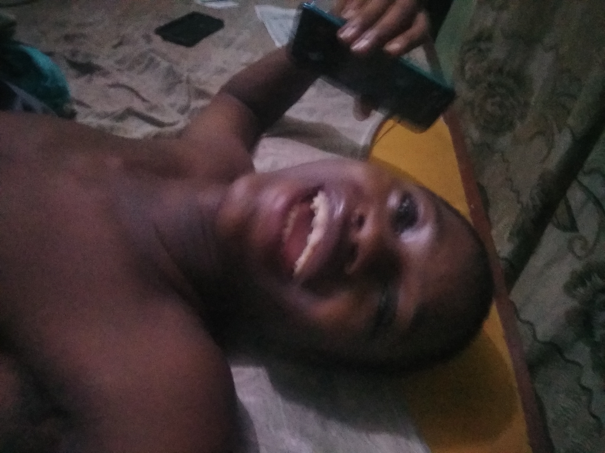 A person is lying on a bed in a room. The person's torso is bare, and they are smiling and looking towards the camera. The person is holding a cell phone above their head. The room appears to be in Shangisha, Nigeria.