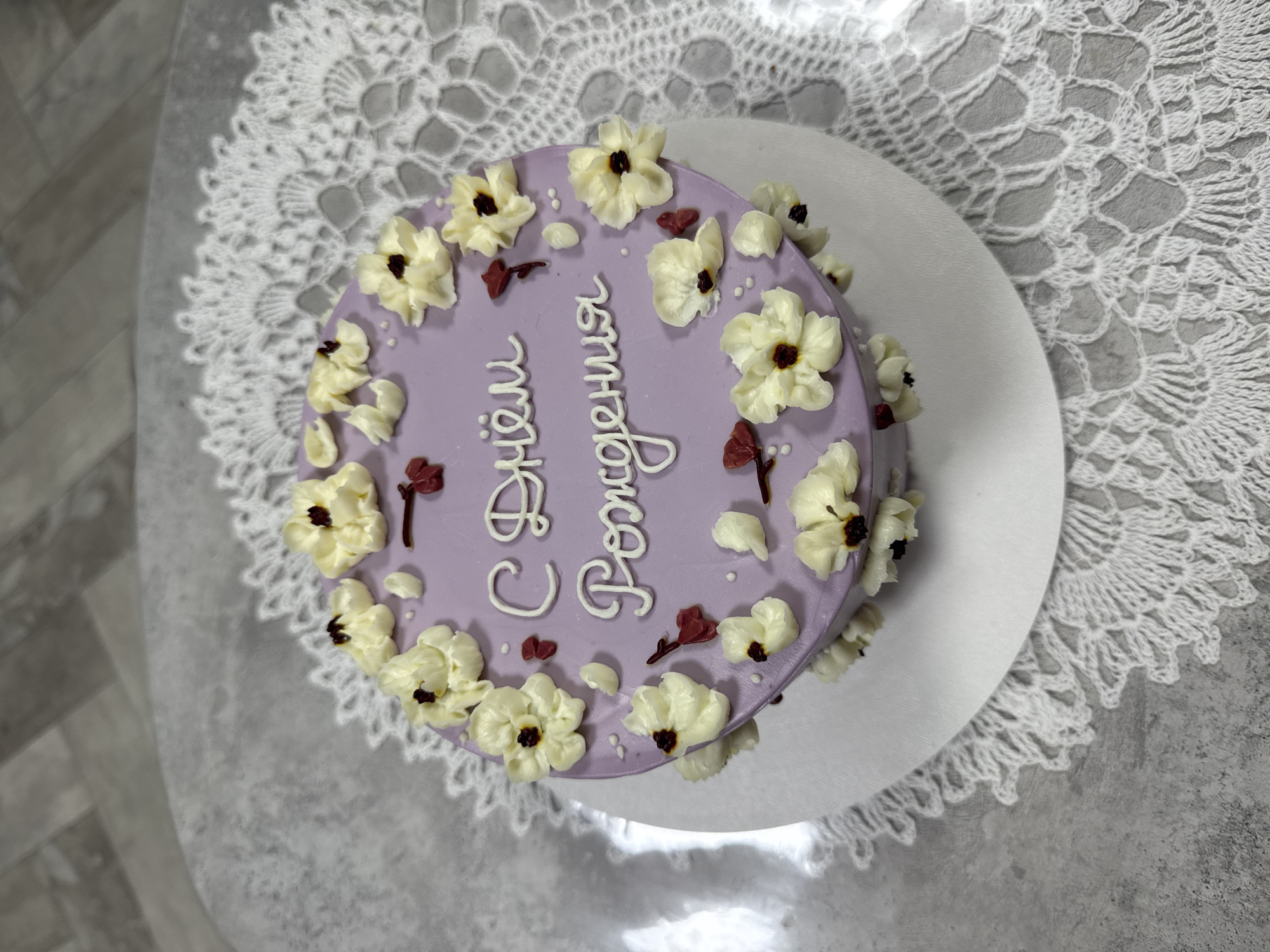 A lilac-frosted cake is displayed on a white doily, which rests on a grey, speckled surface. The cake is adorned with white frosting flowers that have dark centers, and small red frosting accents. White frosting script on the cake reads, "С Днём Рождения" (Happy Birthday). The background shows a grey, wood-patterned floor. There are no people present in the image. The cake is likely located in Sosnoviy Bor, Russia, as indicated by the Cyrillic text on the cake.