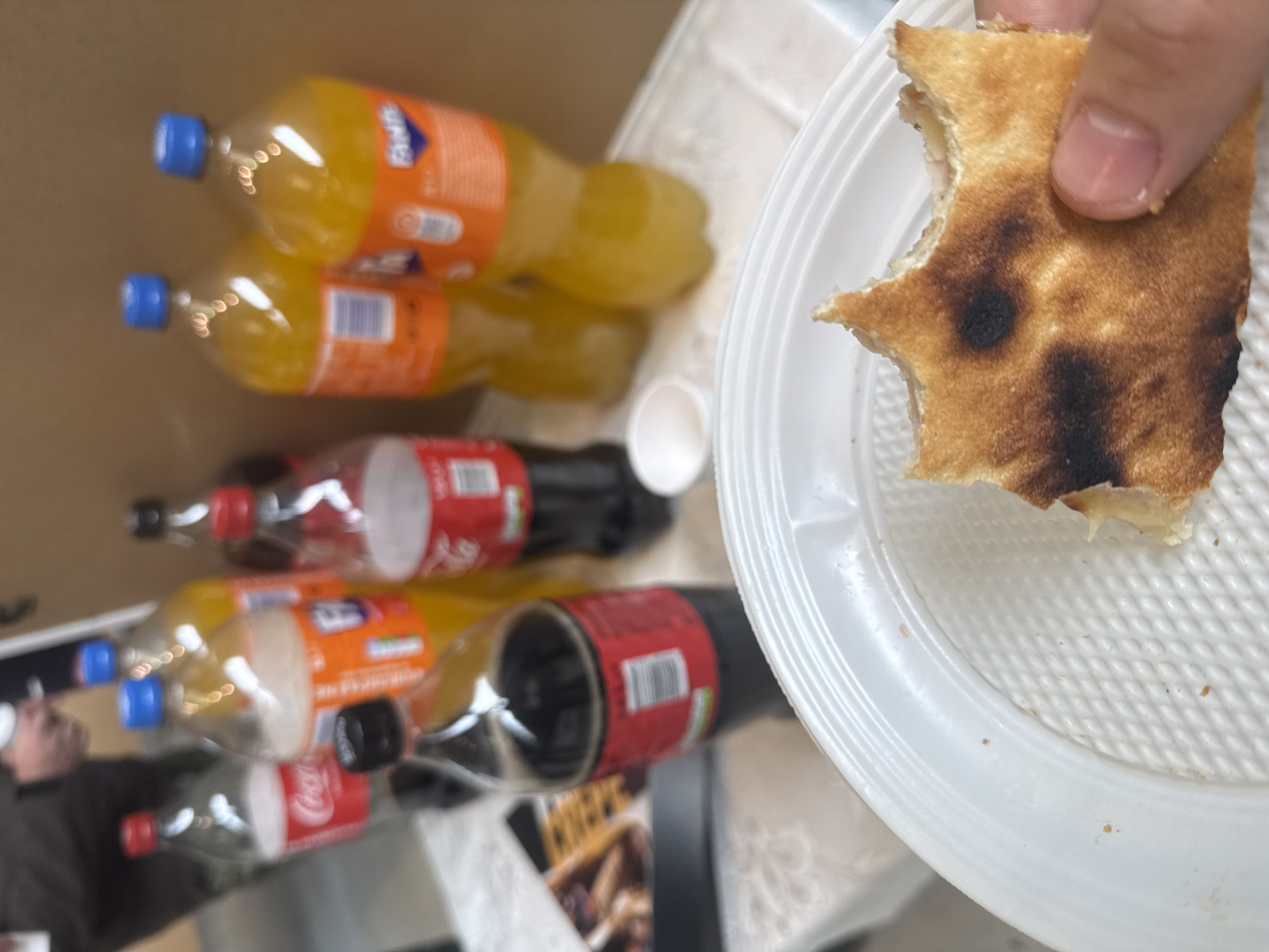 The image shows a variety of items, including several bottles of soda (Fanta and Coca-Cola), a white paper cup, and a white plastic plate. A hand holds a piece of flatbread, possibly pizza, with some burnt spots. The bottles are arranged haphazardly, possibly in a box or bag. The setting appears to be indoors on a table or surface with a white tablecloth. Based on the types of drinks and snacks, the scene could be a casual meal or gathering, but there is nothing in the image to suggest that this is taking place in Sofia, Bulgaria.