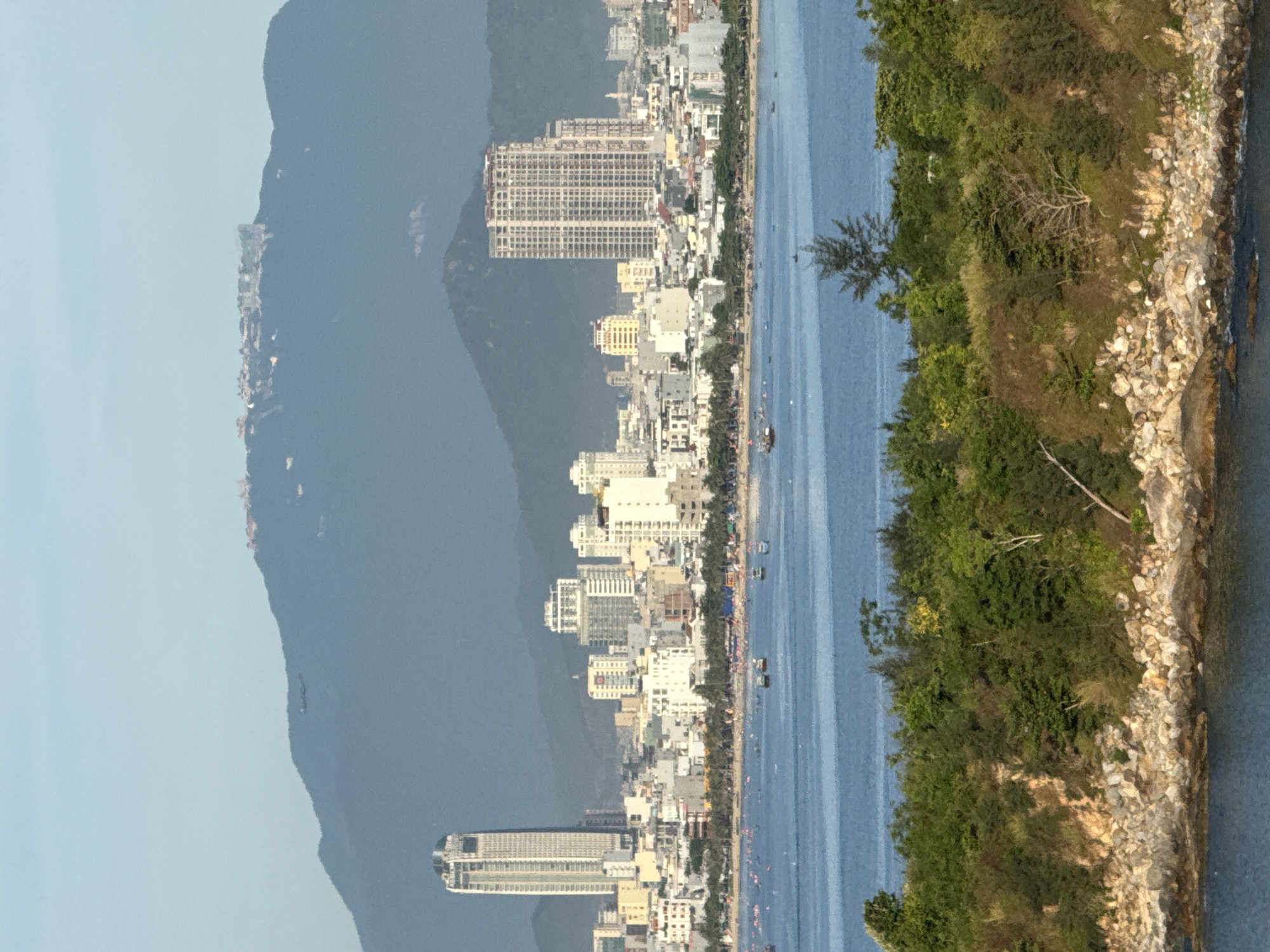 This is a landscape photograph of the cityscape of Da Nang, Vietnam, viewed from a distance across a body of water. In the foreground, a rocky, tree-covered shoreline meets the blue water. Further out, the blue water is dotted with small boats. The middle ground features a densely built urban area with numerous buildings of varying heights, including several prominent high-rise structures. A long, sandy beach runs along the waterfront, with many people visible on the sand and in the water, suggesting a public recreational area. In the background, a large, hazy blue mountain range dominates the horizon. The sky is a pale blue with a slight haze. One of the most notable buildings in the cityscape is a tall, slender white skyscraper with a distinctive patterned facade. Another prominent high-rise is a lighter colored building with many visible floors and balconies. The scene depicts a coastal city with a vibrant urban center set against a natural mountain backdrop.