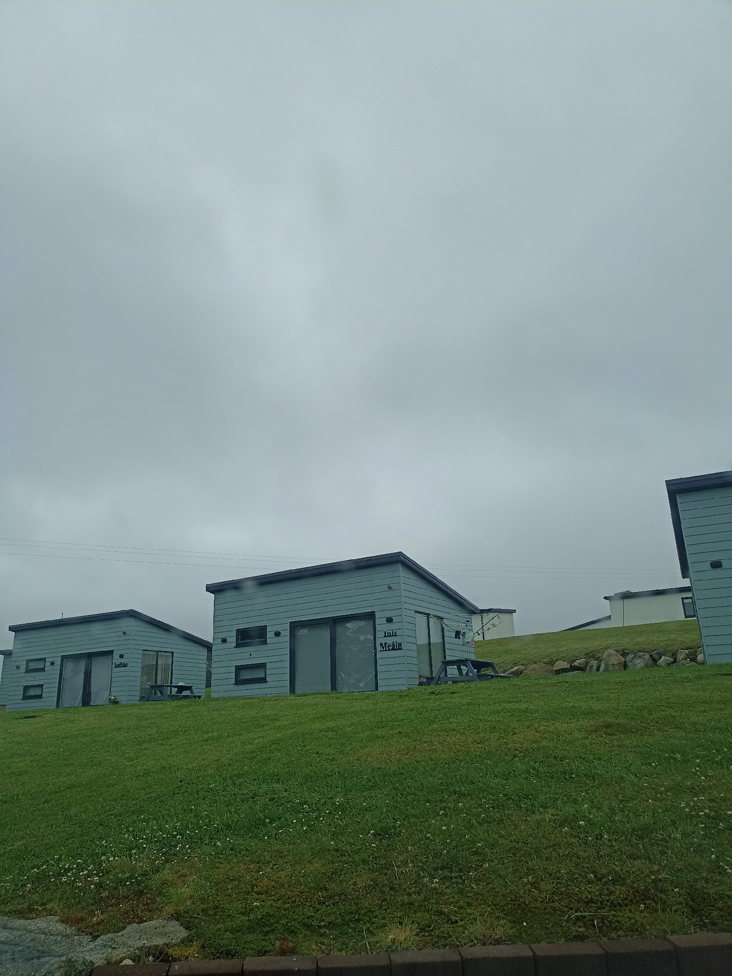 The image shows a cluster of modern, light blue cabins situated on a grassy hillside in Doirí Beaga, Ireland. The cabins have dark gray roofs and dark-framed windows, some of which are large glass doors. One cabin on the left has text on its side that appears to read "Inis Meain". A picnic table is visible in front of this cabin. Further up the hill, more cabins or buildings can be seen, one with white walls and a dark roof. The sky is overcast and gray, suggesting cloudy weather. The foreground consists of a vibrant green lawn dotted with small white flowers. Rocks and a dark border are visible at the very bottom of the image, possibly indicating a pathway or edge of a property. There are no people visible in the image, and the cabins appear unoccupied. The scene is static and depicts a residential or holiday accommodation setting.