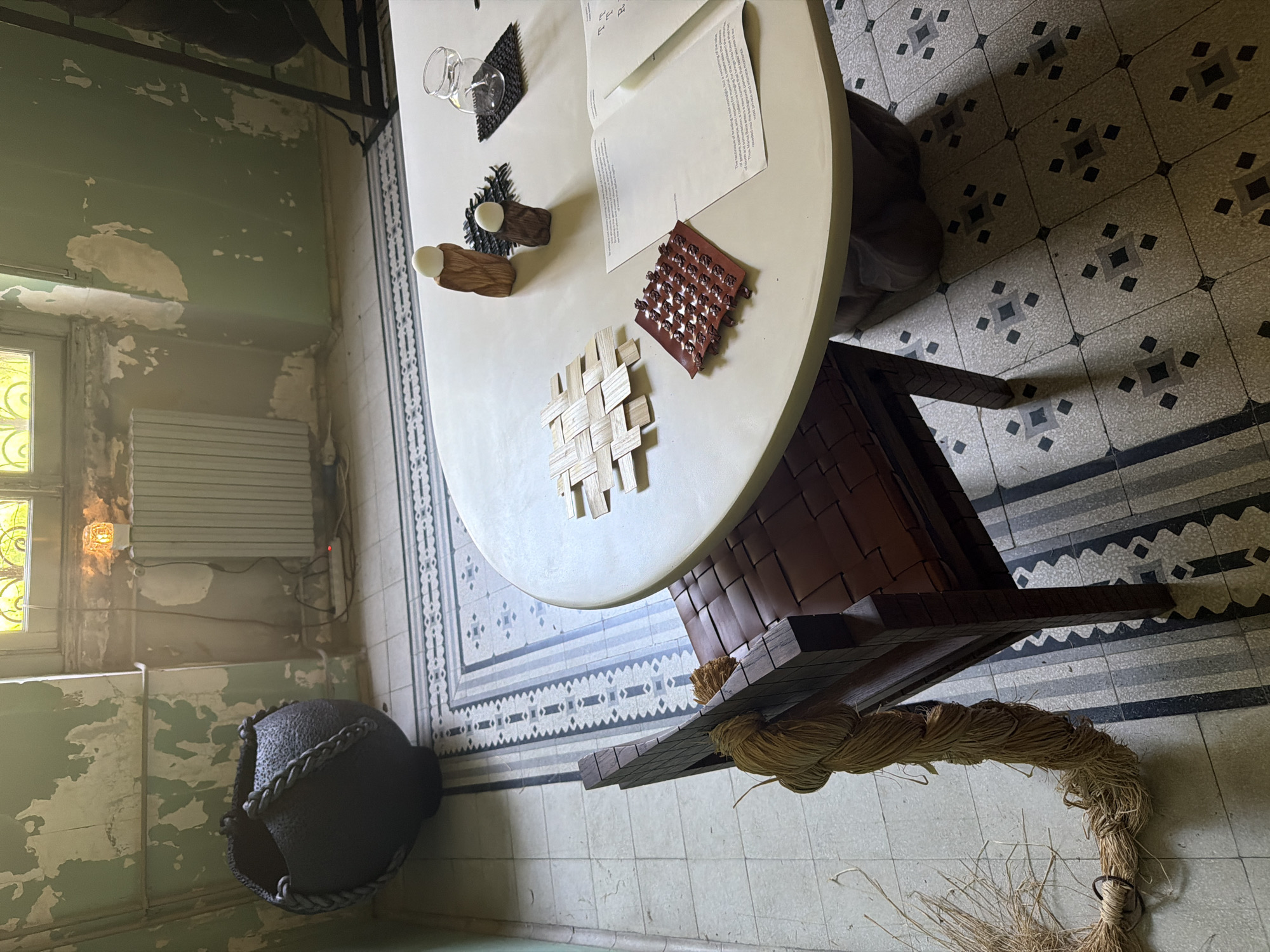 The image depicts an interior scene, likely an art installation, in Milan, Italy. A round table is the central element, displaying various small objects – a glass vessel, textured materials, wooden forms, a woven piece, and printed papers. The table sits atop a unique pedestal-like base, featuring dark wooden elements and rope-like material. The flooring is patterned tile. In the background, walls show signs of age and wear, with peeling paint and exposed plaster. A window is visible, as is a large, dark, spherical object. The overall environment suggests a space with historical character, repurposed for artistic display.