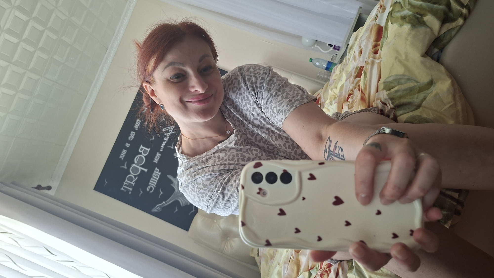 A woman with red hair is taking a selfie in bed. She is wearing a grey leopard-print pajama top and has a tattoo on her left arm and a ring on her left hand. She is holding a white phone case with red hearts. The bed has a yellow and green floral duvet. Behind her, there is a framed piece of art with text on the wall. The room is lit by natural light coming from a window on the left. The scene appears to be a bedroom, likely within a private residence. The location is not specified to be in the city of Myadzyel, Belarus.