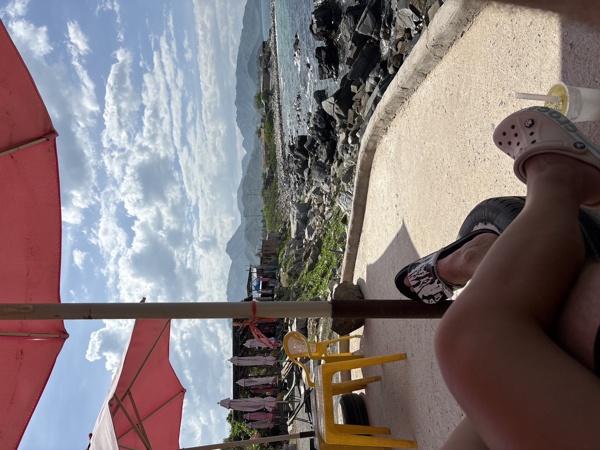 The image captures a sunny day at a coastal location in Vĩnh Lương, Vietnam. The foreground shows a person's legs resting, one with a black and white sandal and the other with a pink sandal. Next to the pink sandal is a clear plastic cup with a straw containing a yellow beverage. In the midground, there is a yellow plastic table and chair set, with several folded pink umbrellas in the background. Beyond the seating area, a rocky coastline meets the ocean, with gentle waves and distant mountains visible under a partly cloudy sky. A few buildings and vegetation are scattered along the coast. The scene appears to be an outdoor seating area at a beachside establishment.