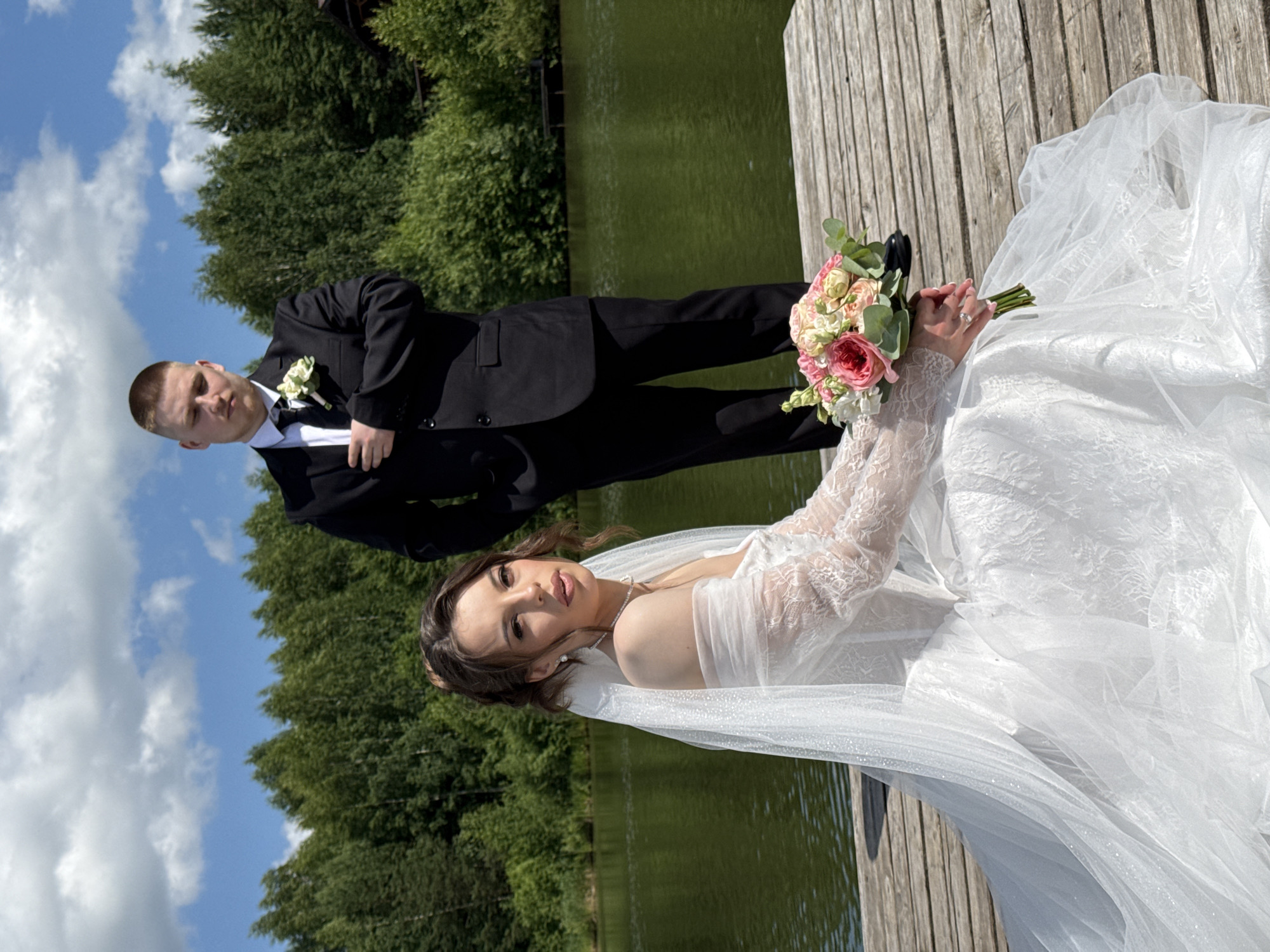 The image captures a wedding scene set on a wooden pier next to a body of water. The subjects are a bride and groom, dressed in wedding attire. The groom is wearing a black suit with a white shirt and a dark tie, adorned with a white boutonniere. He is looking down with a serious expression. The bride is in a white wedding dress with lace sleeves and a veil. She is holding a bouquet of pink and white flowers and is looking towards the camera with a gentle expression. The setting appears to be outdoors, with lush green trees and a bright blue sky with white clouds visible in the background. The water is a calm green color. The lighting suggests it is daytime, likely sunny. The overall mood is romantic and celebratory. There is no visible text in the image. The location context is Votkinsk, Russia.