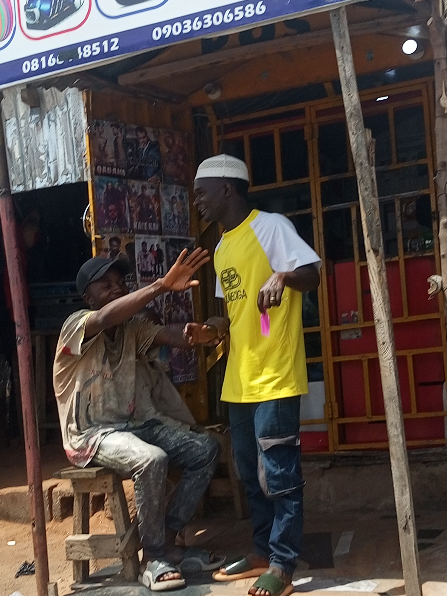 In Gombe, Nigeria, two men stand in front of a shop displaying movie posters. The man on the left, seated on a wooden stool, wears a dirty, paint-splattered light-colored shirt and dark pants, a black baseball cap, and sandals. He is gesturing with his right hand. The man on the right, standing, wears a white and yellow t-shirt with a logo, blue jeans, and sandals. He is holding a pink object in his right hand. The shop's facade has a yellow door with red panels. A sign with phone numbers is visible above the shop. The ground is dusty and uneven.