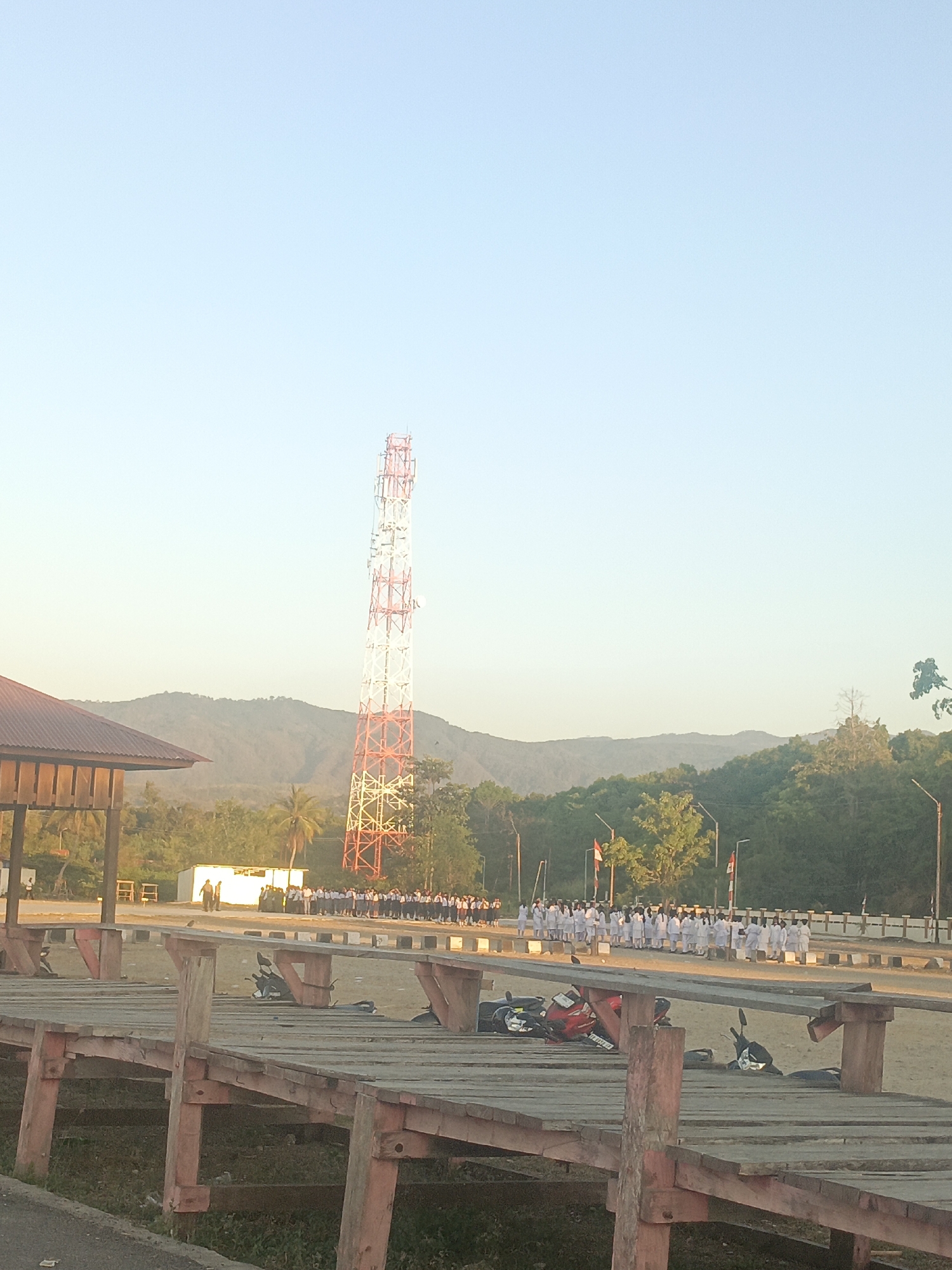 In Lewoleba, Indonesia, a group of individuals in uniform stand in formation. A tall, red and white telecommunications tower dominates the background. In the foreground, a wooden structure with weathered planks and red-painted supports runs horizontally across the image. Several motorcycles are parked or lying on the ground near the wooden structure. The scene is set against a backdrop of rolling green hills and a clear, pale blue sky, suggesting it is either early morning or late afternoon. The overall atmosphere is serene and orderly.