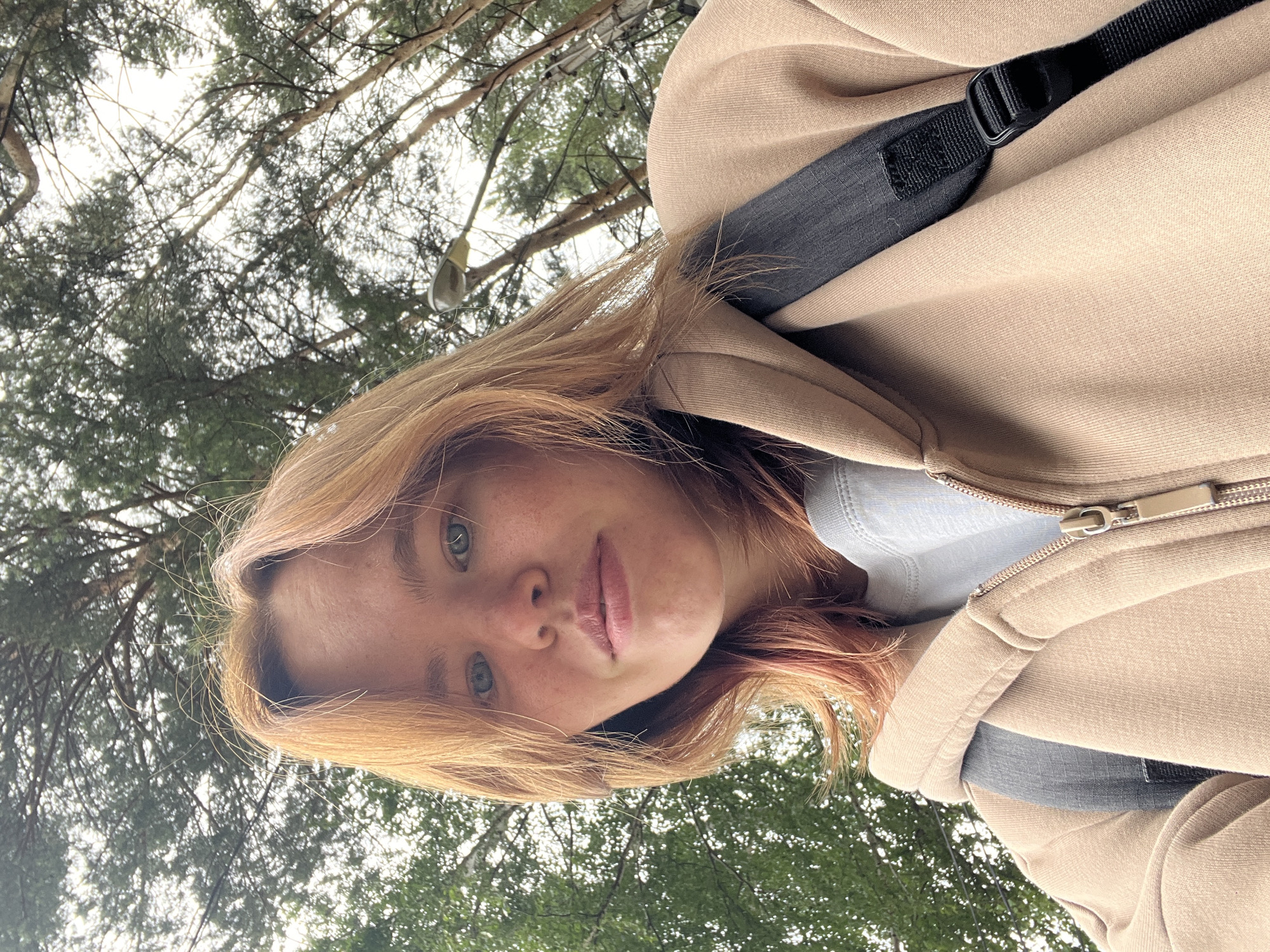 The image is a selfie taken from a low angle looking up at a young woman with blonde hair, blue eyes, and fair skin. She is wearing a light blue t-shirt under a tan hooded jacket, with a black backpack strap visible over her shoulder. The background consists of trees with green foliage and a cloudy sky, suggesting an outdoor setting, possibly a park or wooded area within the city of Chaikovsky, Russia, given the provided context. A street lamp is also visible among the trees. The woman is looking directly at the camera with a neutral expression.