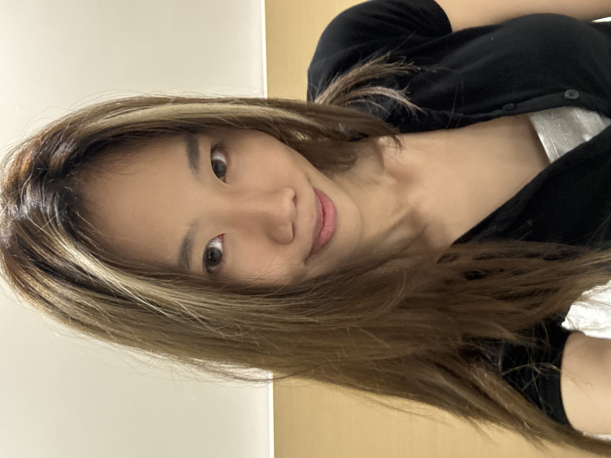 A young Asian woman is facing the camera. She has brown hair with blonde highlights. She is wearing a black button-down shirt and a white undershirt. The background consists of a light beige wall with a darker beige trim along the bottom. The photo appears to have been taken indoors, possibly in a room with artificial lighting. There is no information to suggest this photo was taken in Yuzhong District, China.