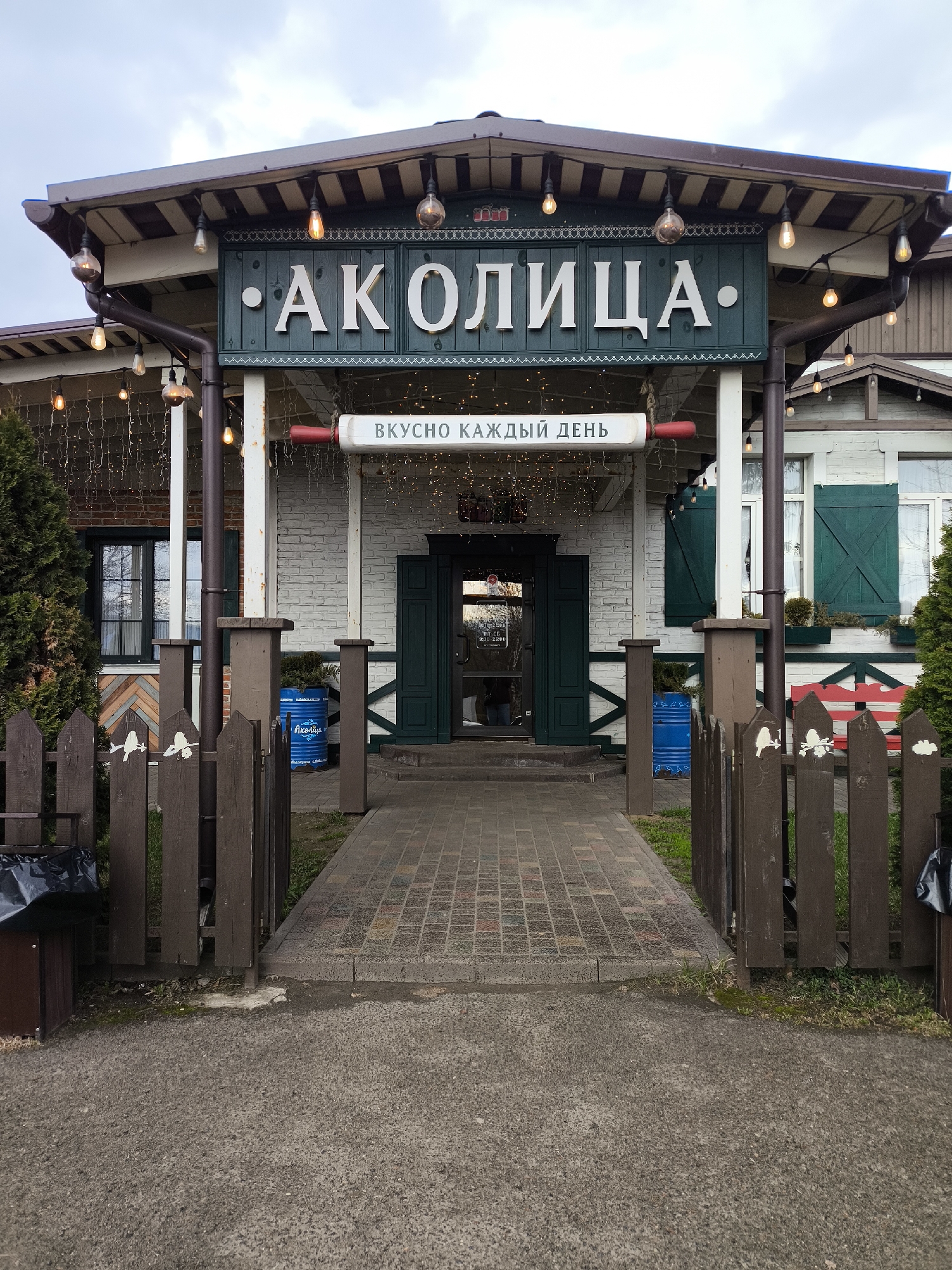 This image captures the exterior of a charming building, likely a restaurant or cafe, under an overcast sky. The establishment, named "• АКОЛИЦА •" (Akolitsa), is fronted by an inviting porch-like structure with an overhanging roof adorned with decorative string lights.
The main building features white brick walls and dark green double doors at the central entrance. Windows on either side are fitted with matching dark green louvered shutters. A paved brick walkway leads directly to the entrance, flanked by sturdy white support pillars and a dark brown wooden picket fence with decorative cut-outs. On the left, a tall evergreen bush adds greenery, while on the right, a red and white striped wooden bench provides a seating area. Two blue industrial-style barrels, one clearly marked with "АКОЛИЦА" in white Cyrillic letters, are positioned as decorative elements near the entrance.
Above the entrance, a prominent dark teal sign displays the establishment's name, "• АКОЛИЦА •", in large white Cyrillic letters. Beneath it, a white banner proclaims "ВКУСНО КАЖДЫЙ ДЕНЬ" (Delicious Every Day), accompanied by a red decorative piece resembling a rolling pin. A small sign on the door indicates operating hours, partially visible as "9:00 - 23:00".
No people are visible, but the well-maintained appearance and visible signage suggest an active business. The ground in the foreground is asphalt, transitioning to the brick path. The overall impression is one of a cozy, rustic, and welcoming dining spot, consistent with its location in Andreikovo, Russia.