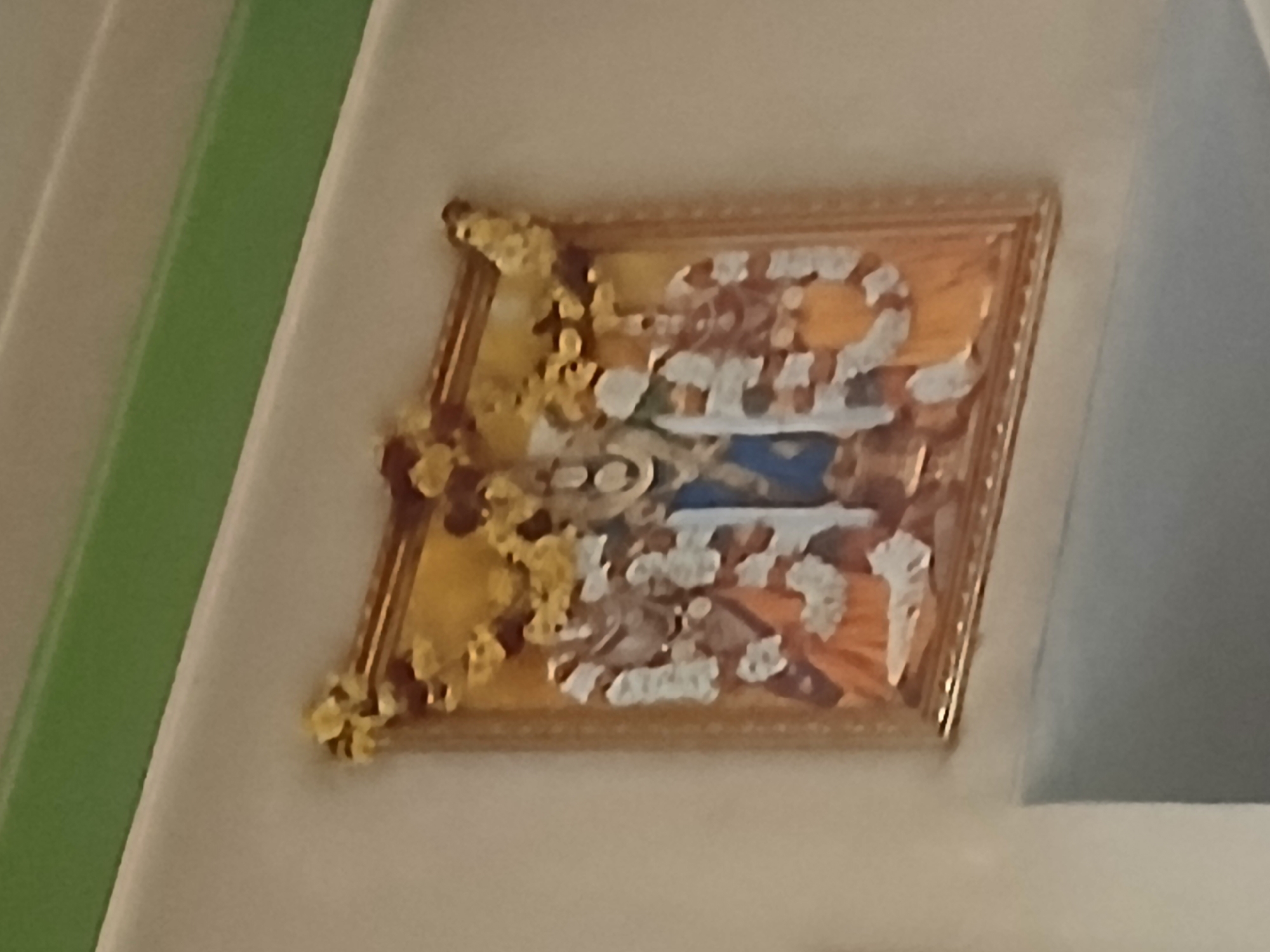A framed painting hangs on a wall. The painting has a gold frame. The scene is indoors, likely in Bengaluru, India, based on the style of art depicted, although specific location cannot be verified from the image alone. The painting depicts human figures and possibly animals, though obscured by wear or decorative elements.