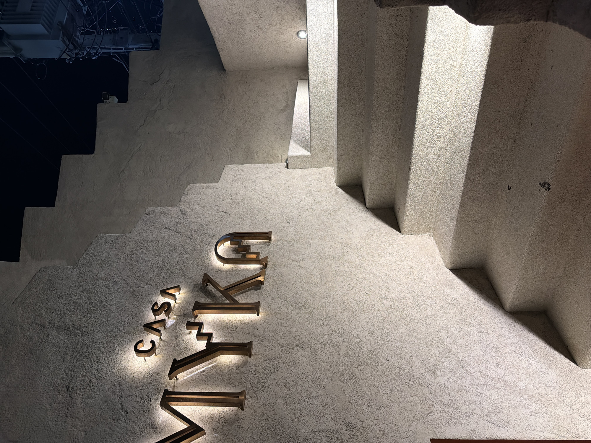The image presents an architectural detail, likely of a building exterior, in Pattaya City, Thailand. The focus is on a section of a textured white or off-white wall adjacent to a staircase or tiered architectural element. The steps are also finished in the same textured material. The letters "CASA VYKU" are vertically oriented on the wall, with lighting effects accentuating each letter. The lighting suggests nighttime or low-light conditions. Overhead, electrical wires are visible. The overall aesthetic is modern and minimalist.