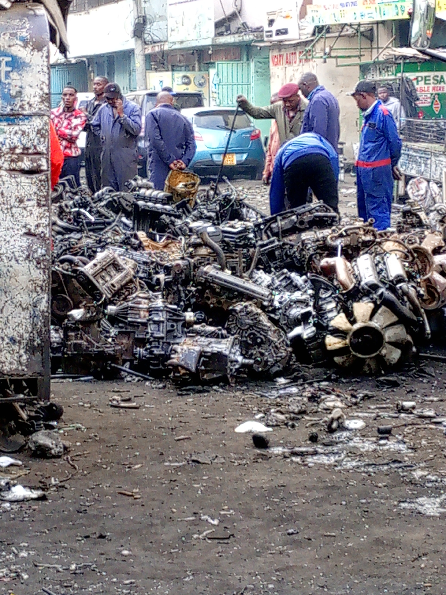 The image captures a bustling scene in Nairobi, Kenya. A large pile of used car engines and parts dominates the foreground, suggesting a junkyard or auto repair shop. Several men are gathered around the pile, some seemingly inspecting or sorting through the parts. Their clothing varies, ranging from casual attire to work overalls. In the background, a blue car is parked along a street lined with buildings. One building has a sign that reads "NCATY AUTO SPARE PARTS". The ground appears muddy and strewn with debris, indicating a working environment. A sign on another building advertises "PESA ABLE HERE." The overall atmosphere suggests a busy, gritty urban setting centered around the automotive industry.