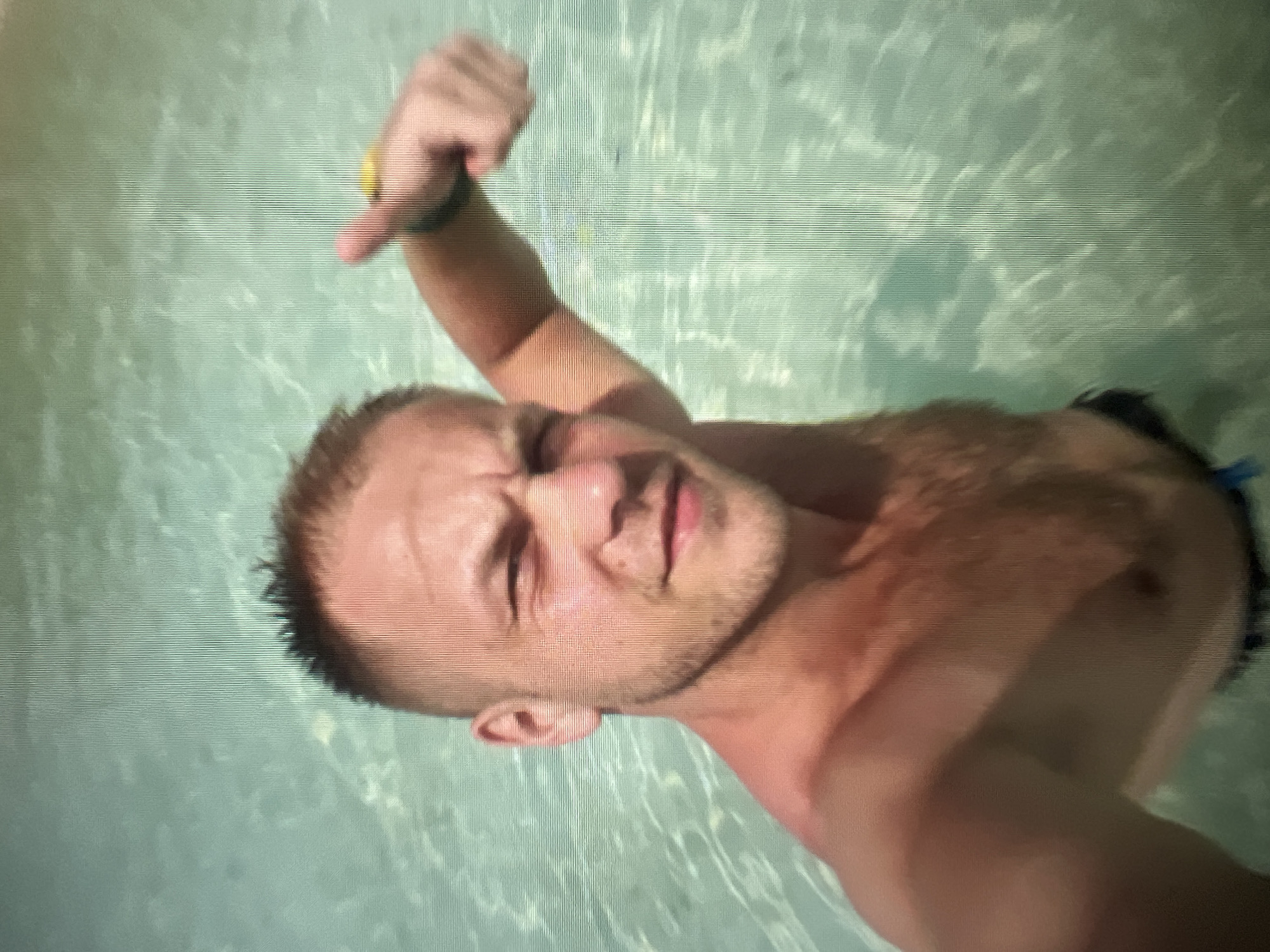 A man is in the water, giving a thumbs up to the camera. He is shirtless and has a short haircut. The water is clear and appears to be a pool or shallow ocean. The man is wearing a wristband on his left arm. The image quality is poor and pixelated, making it difficult to discern fine details. There is no indication of the location being the city of Qarağandı city administration, Kazakhstan.