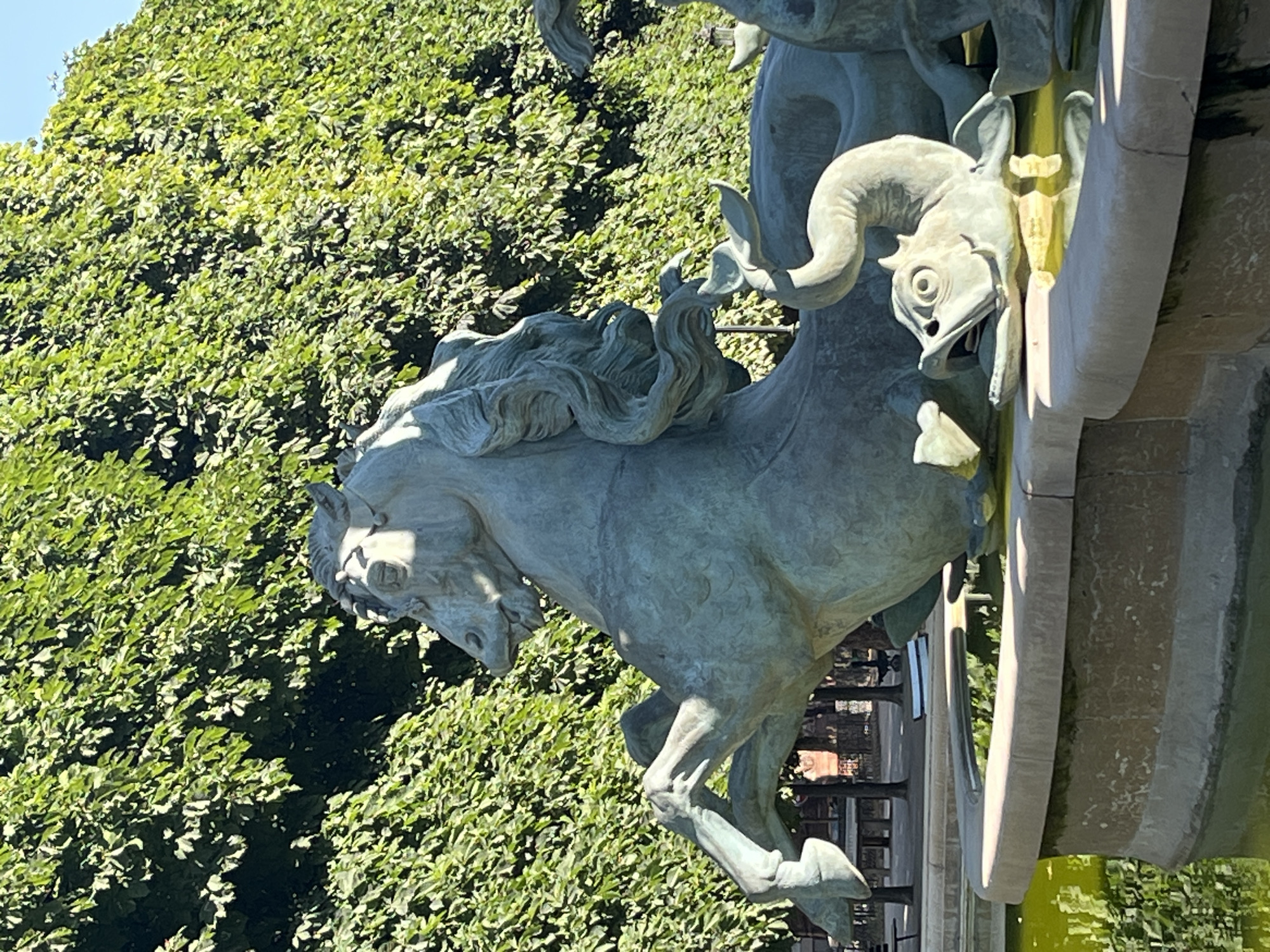 The image shows a bronze sculpture of a horse, part of a fountain, in Paris, France. The sculpture is detailed, with the horse rearing up, its mane flowing. Below the horse, a sculpted fish with an open mouth appears to be spouting water, though no water is visible in the image. The fountain is surrounded by a stone basin filled with murky green water, reflecting the lush, green foliage behind it. The foliage appears to be a dense hedge or row of trees, creating a natural backdrop. In the background, through the foliage, there are glimpses of park benches and possibly other park structures, suggesting the sculpture is located in a public garden or park. The lighting indicates it is daytime, with sunlight casting shadows and highlighting the metallic sheen of the sculpture.