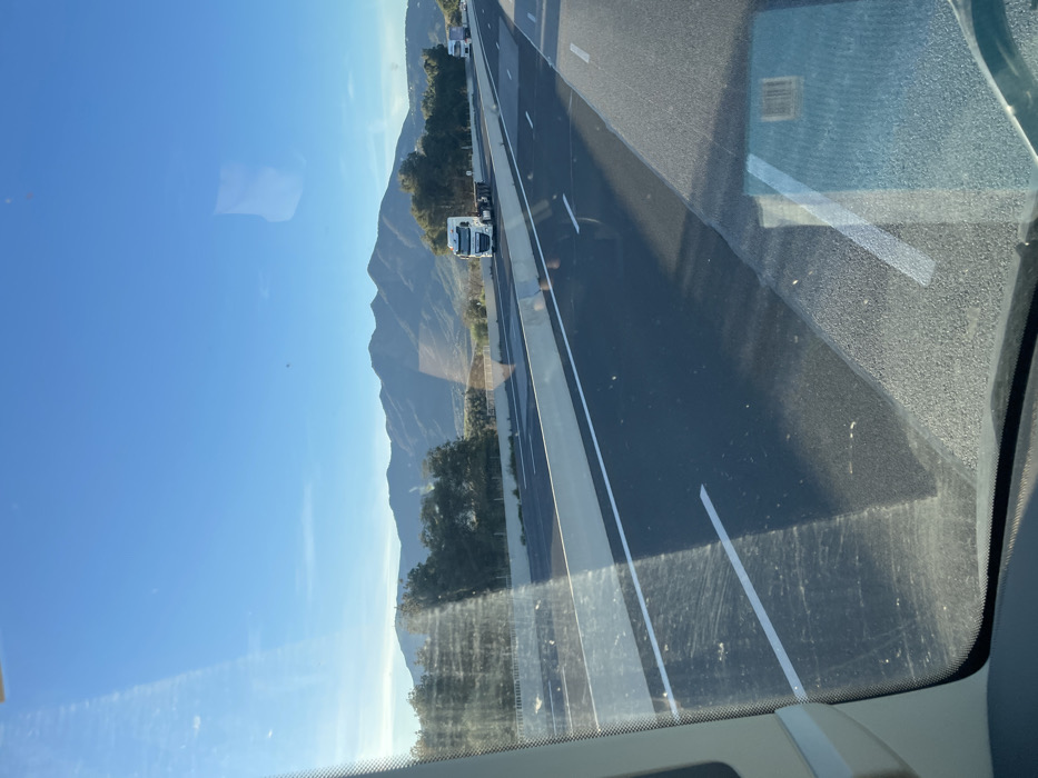 The image captures a daytime highway scene, viewed from inside a vehicle through its windshield, situated near Tresserre, France. The upper portion of the frame is dominated by a clear, bright blue sky with minimal cloud cover. A range of dark green, rugged mountains stretches across the horizon, with sporadic trees visible in the mid-ground between the mountains and the roadway. The foreground features a multi-lane highway constructed of dark asphalt, delineated by white dashed and solid lane markings. A light grey concrete median barrier separates the directions of traffic, casting distinct shadows onto the road surface, indicative of strong, direct sunlight. In the lanes of opposing traffic, a white DAF semi-truck tractor, without a trailer, is visible in the leftmost lane, traveling away from the perspective of the camera. Further down the opposing road, another white semi-truck is partially discernible. The vehicle's windshield, framing the lower-left portion of the image, exhibits reflections and minor surface imperfections.