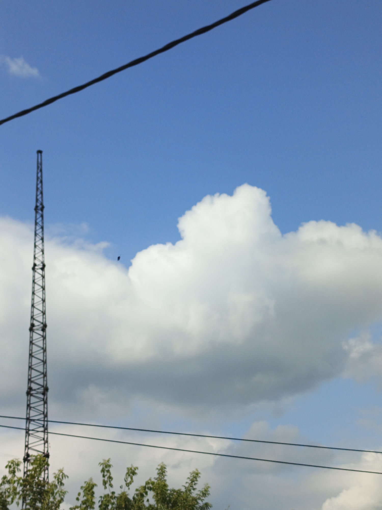A tall, thin, lattice radio tower stands to the left of frame. The tower is silhouetted against a bright blue sky with large, fluffy white clouds. A thin, black wire stretches diagonally across the upper portion of the frame, originating from the top left corner and extending towards the top right. Another black wire runs horizontally across the lower portion of the frame. Below the tower and wires, the tops of green trees are visible. A small, dark, kite-like object is visible in the sky between the radio tower and the clouds. The scene appears to be an outdoor shot on a sunny day. The image does not depict any people or specific city landmarks that would allow for precise identification of the location within Zlatoust, Russia.