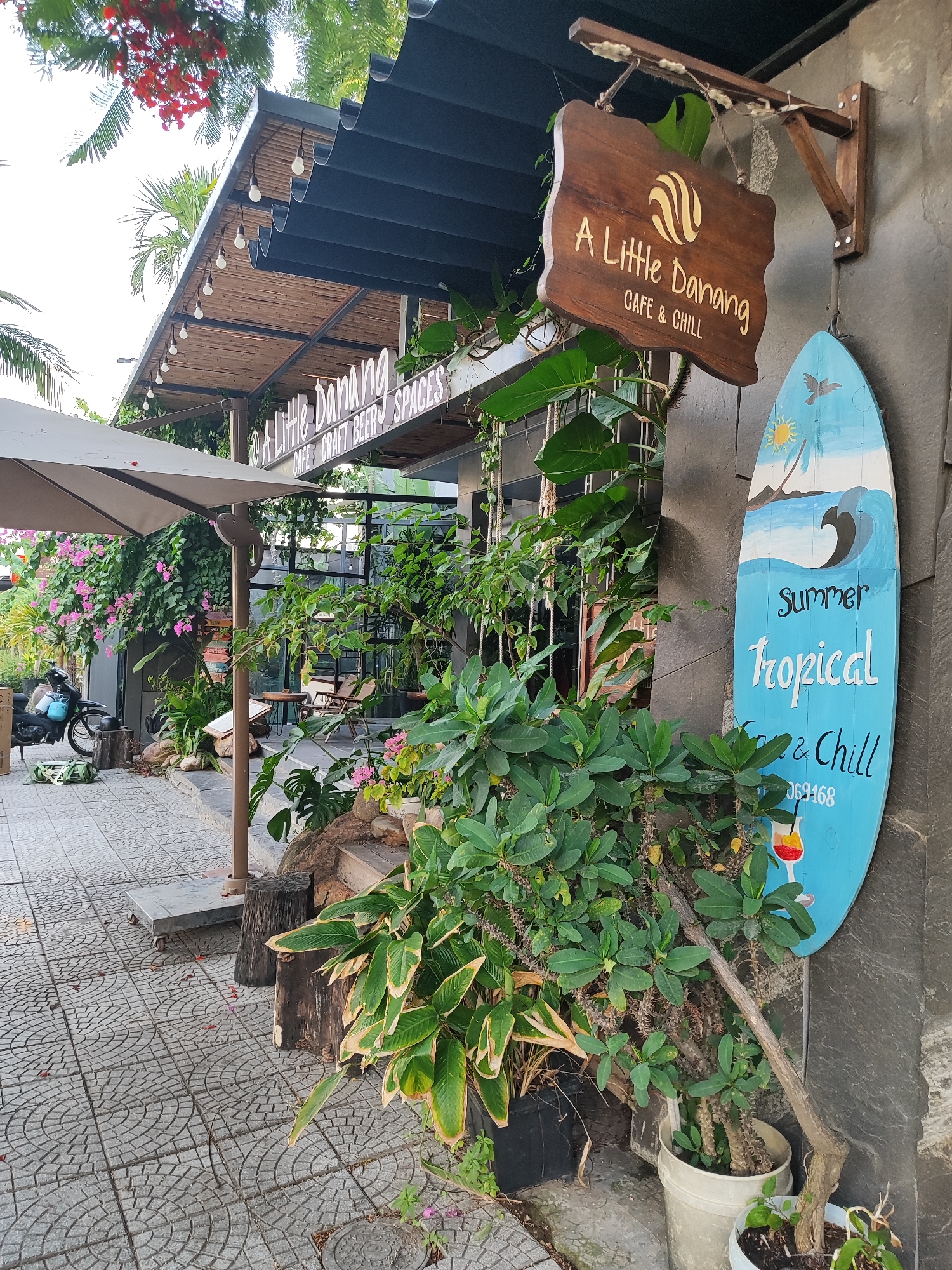 The image shows the exterior of a cafe and chill establishment in Da Nang, Vietnam. A wooden sign hanging from a bracket displays "A Little Danang CAFE & CHILL" with a circular logo. To the right of this sign, a blue surfboard-shaped board with painted waves, sun, and a bird reads "Summer Tropical Cafe & Chill" and a phone number, "069168," and a cocktail illustration at the bottom. Lush green plants and flowers surround the cafe's entrance, with a motorcycle parked to the left. The cafe has an outdoor seating area with tables and chairs visible through glass doors. The pavement in front of the cafe is tiled in a geometric pattern. The overall scene suggests a relaxed, tropical atmosphere.