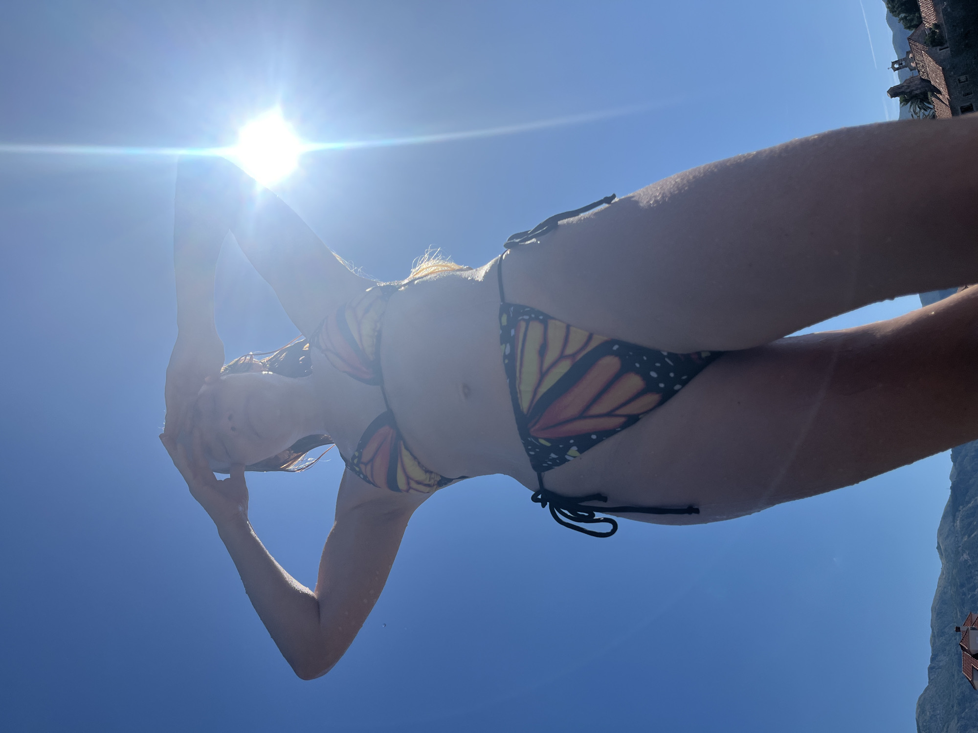 The image shows a person in a bikini with a monarch butterfly pattern, taken from a low angle looking up at the sky. The sun is bright and directly overhead, creating lens flare. The person's arms are raised above their head, and they appear to be holding something or shielding their face from the sun. The background is a clear blue sky. On the right side of the image, there are distant buildings and a mountain, suggesting an outdoor location, possibly a coastal area. The overall impression is one of a sunny day, likely during the daytime. The image appears to be taken in Budva, Montenegro, based on the provided location context. No visible text is present in the image.