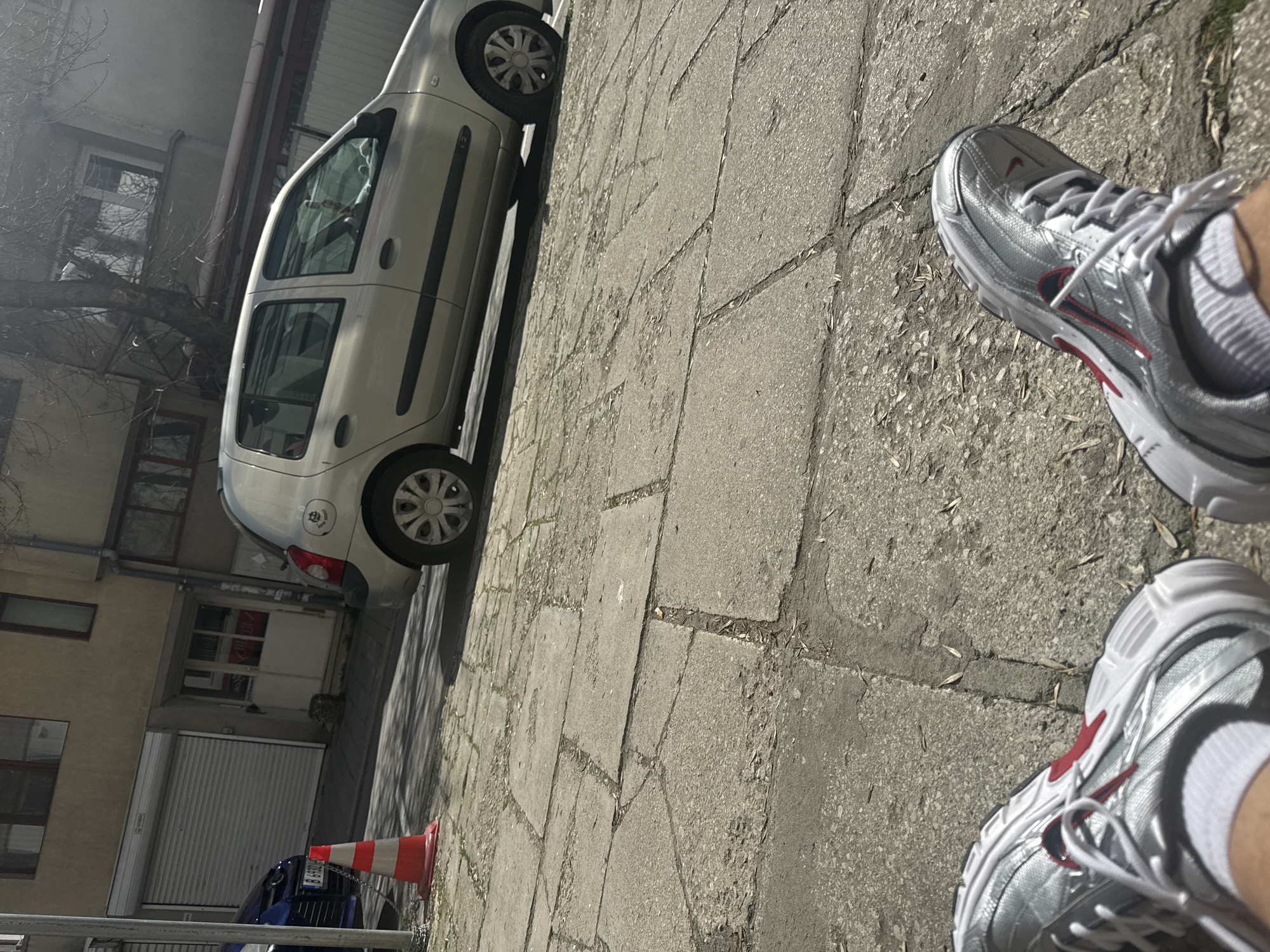 The image shows a first-person view of a person standing on a paved sidewalk next to a silver car. The person is wearing silver and white sneakers with red accents and white socks. In the background, there is a building with a closed roller shutter. An orange and white traffic cone is present near a blue car. The image was taken in Varna, Bulgaria.
