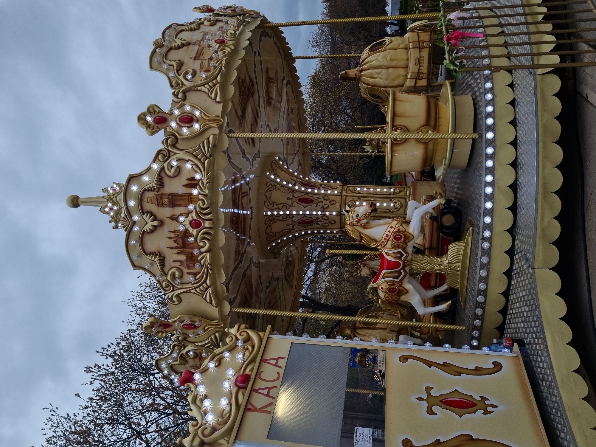 The image shows a carousel in Sofia, Bulgaria. The carousel is decorated with images of London landmarks, including Big Ben. There are horses and other animals for people to ride on. The carousel has a decorative canopy with lights. The sky is overcast. The words "KACA" and "РАБОТНО ВРЕМЕ" (working hours) are visible on a sign. The carousel is in a park-like setting with trees in the background.