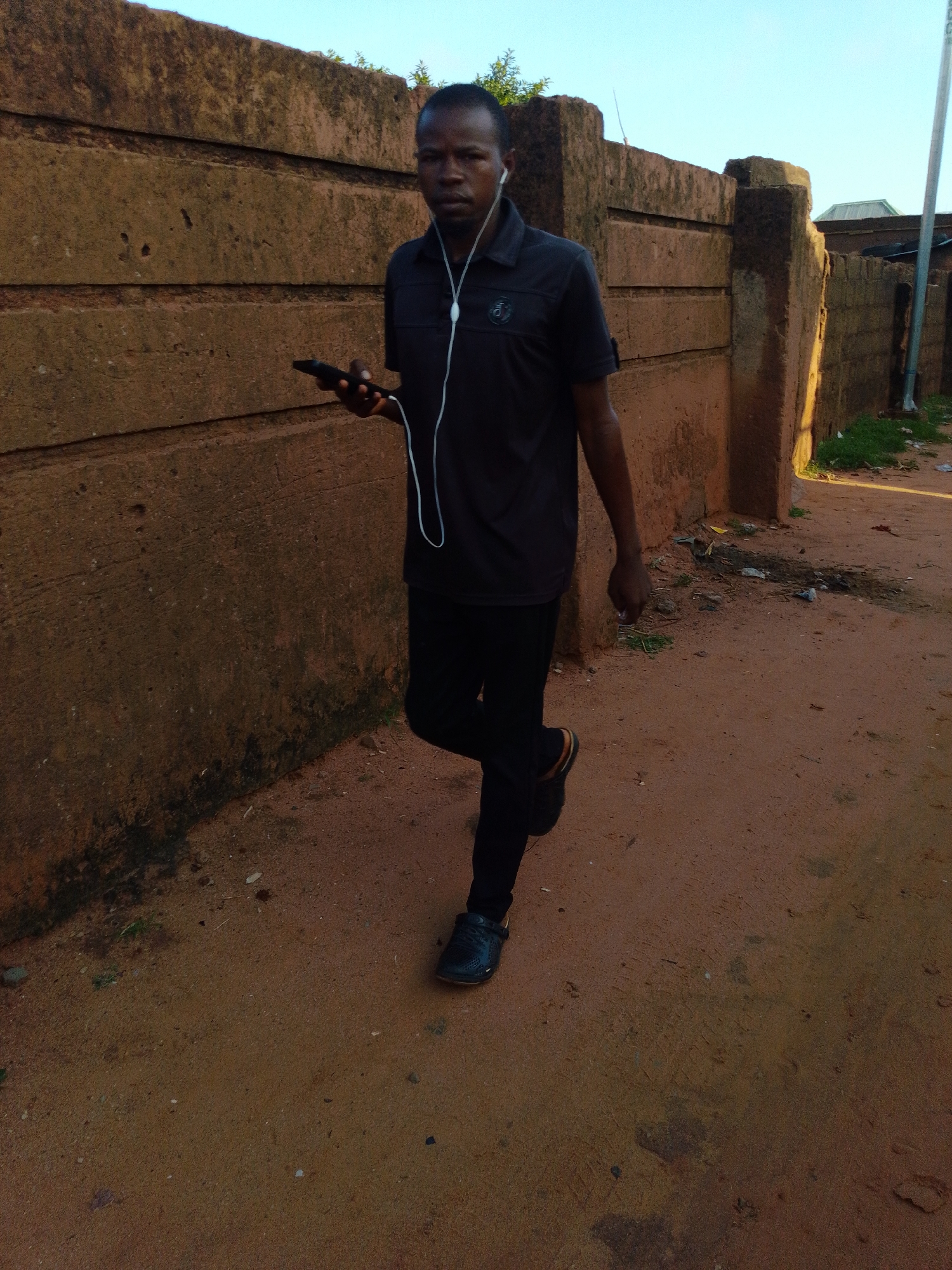 A Black man is walking in front of a textured, reddish-brown wall in Tempure, Nigeria. He is wearing a dark polo shirt, black pants, and dark shoes. He has white earbuds in his ears and is holding a black smartphone in his right hand. The ground is a sandy, reddish-brown dirt path. There is some scattered debris and greenery visible on the ground and along the base of the wall. The sky is a clear light blue.
