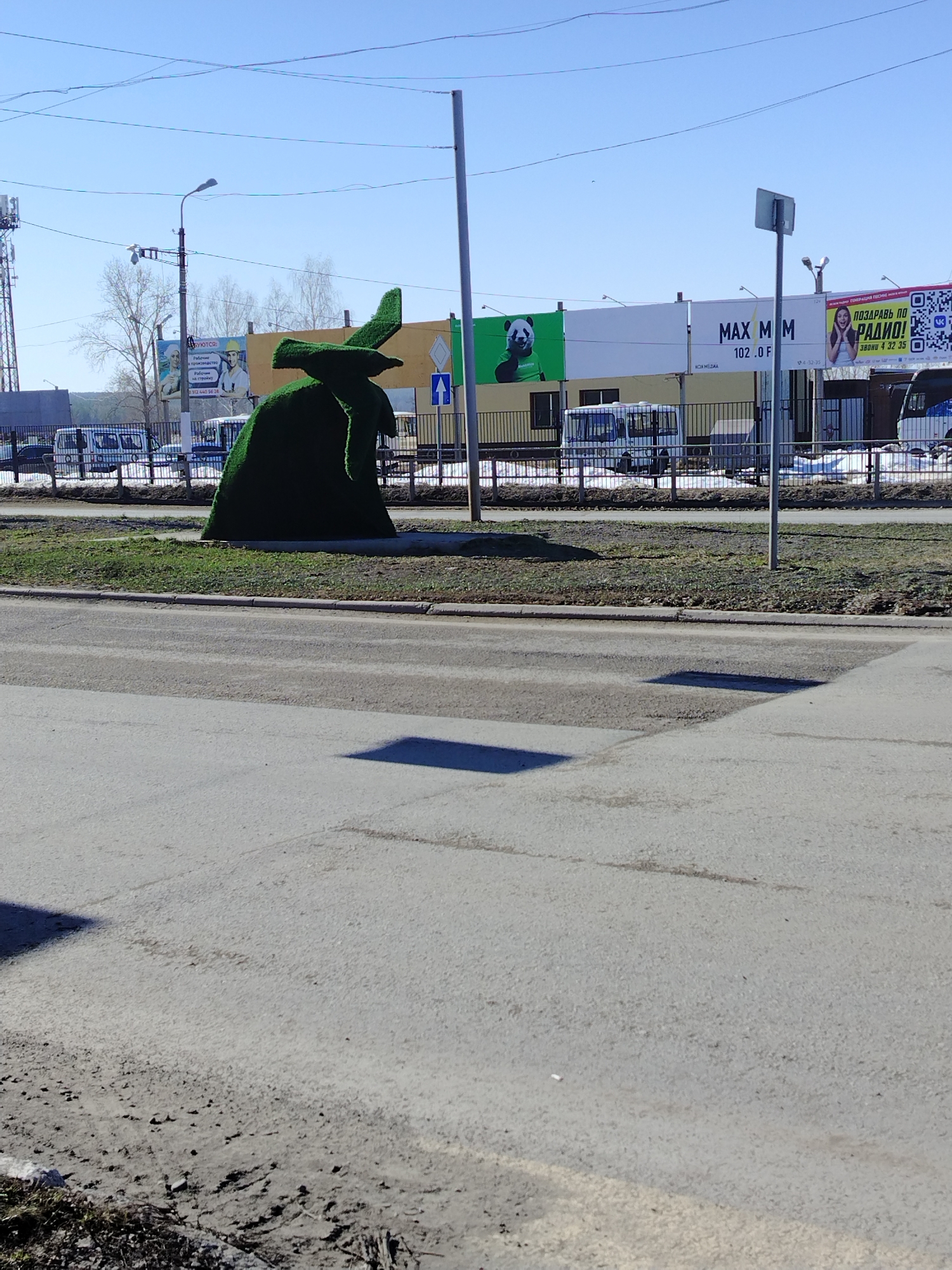 The image shows a green topiary sculpture, possibly representing a person with a hat, located in Chaikovsky, Russia. The sculpture sits on a grassy traffic island bordered by a curb and a four-lane asphalt road with shadows. Behind the island, a black metal fence separates the road from a row of buildings with large advertising banners. The banners advertise various products or services. Some are generic, and one shows a panda. Street lamps and a communication tower are visible in the background, under a clear blue sky. The image captures a typical urban street scene on a sunny day.
