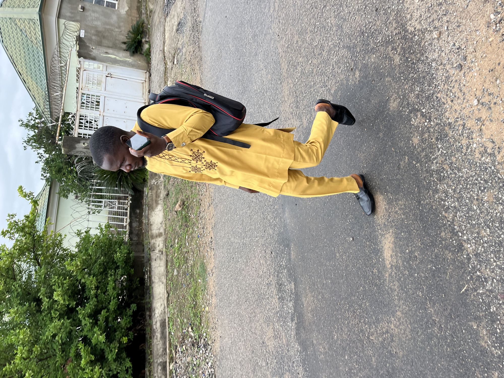 A man is depicted walking outdoors on an asphalt road in Abuja, Nigeria, while looking at his smartphone. He is dressed in a vibrant yellow traditional outfit, possibly a kaftan or dashiki, featuring intricate brown embroidery around the neckline and chest. He wears black loafers on his feet and carries a black backpack on his shoulders. A watch is visible on his left wrist.
The man is captured mid-stride, with his head tilted downwards, focused on the screen of his phone which he holds in his right hand. The setting appears to be a residential area. To the left of the man is a building with a light green pitched roof and light-colored walls. A white metal gate, topped with coils of razor wire for security, provides access to the property. Lush green trees and bushes line the street and are visible behind the gate and building.
The ground is a textured asphalt road surface, speckled with lighter-colored gravel and patches of dirt. A concrete curb or sidewalk edge is visible on the far left. The sky appears overcast, suggesting a cloudy day. There is no discernible text visible in the image. The overall impression is of a casual moment during the daytime.