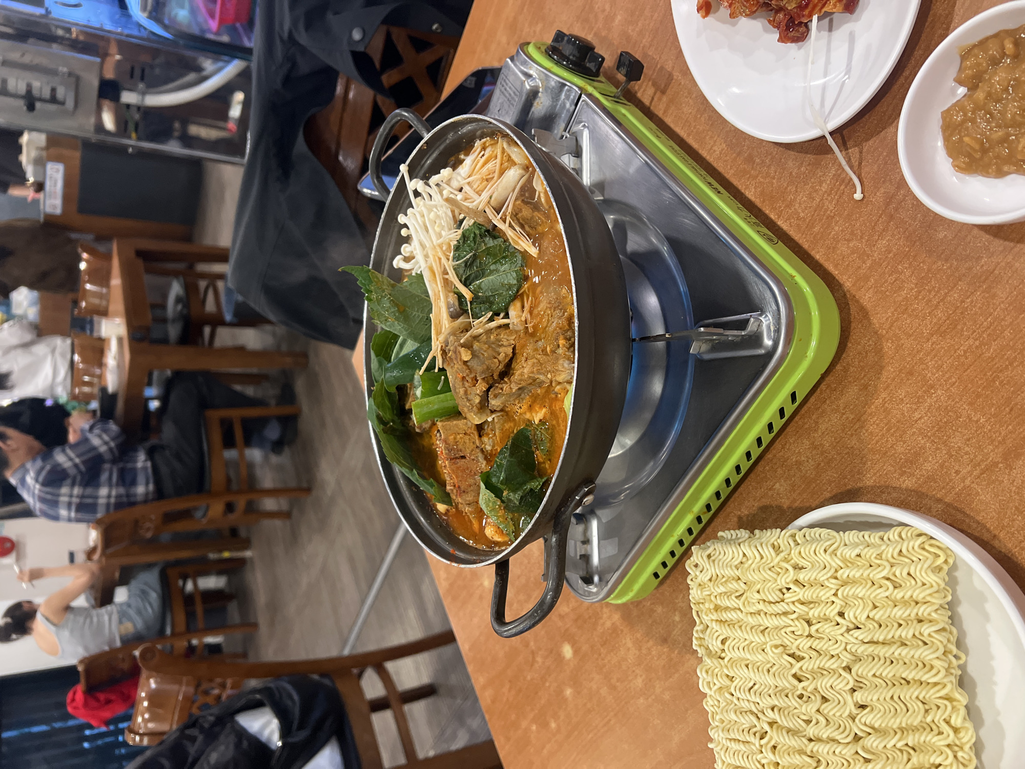 **Scene:** A table in a restaurant in Seoul, South Korea, is set with a Korean stew simmering on a portable gas stove. The stew contains meat, vegetables, and mushrooms. Alongside the stew are plates of banchan (side dishes), including a white plate of what appears to be ramen noodles, and a small dish of sauce.
**People:** Several blurred individuals are seated at the table and in the background, suggesting a busy dining atmosphere.
**Objects:** The stew is in a black pot with handles, placed on a green and silver portable gas stove. White ceramic dishes hold various side dishes. The table is wooden, and the background features restaurant furniture like wooden chairs and possibly a fish tank.
**Actions:** The stew is actively simmering on the stove, indicating cooking and consumption. People in the background are seated and likely eating.
**Interactions:** The food arrangement suggests a communal dining experience, typical of Korean meals.
**Environment:** The restaurant appears moderately busy, with standard dining furniture. The presence of the gas stove indicates an interactive cooking experience at the table.