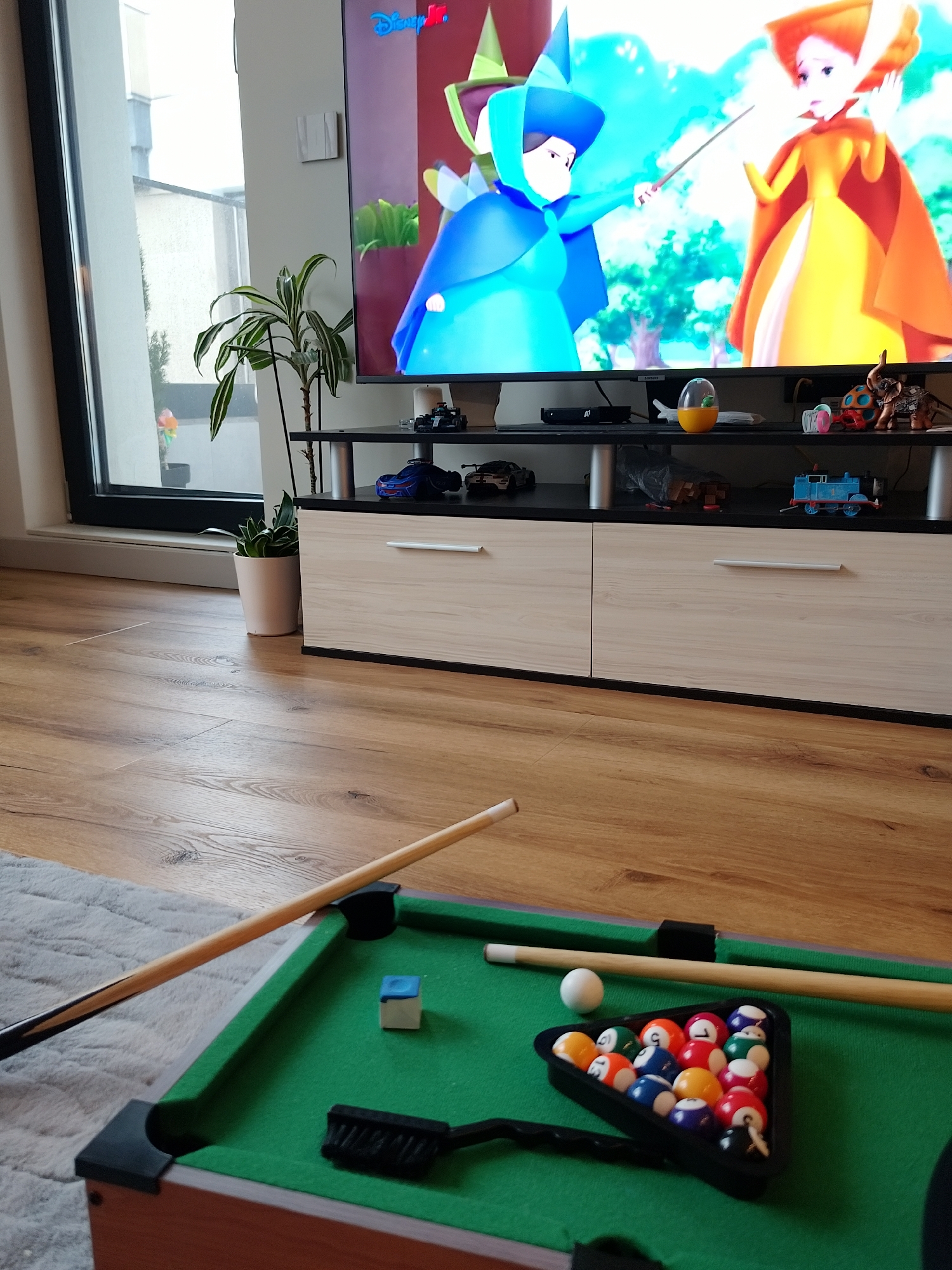 The image presents a bright indoor scene, likely a living room in Sofia, Bulgaria, centered around a large television and a miniature pool table. A large flat-screen TV displays an animated cartoon, with the "Disney Jr." logo visible in the top left corner. The animated scene features two female characters in fantastical attire; one in blue holds a magic wand, pointing it towards the other character dressed in yellow and orange.
Below the television is a black and light wood-grain entertainment unit. Its top shelf is adorned with various objects, including toy cars (a blue race car and a black car), a blue Thomas the Tank Engine toy, a yellow egg-shaped item, an elephant figurine, and a device marked with the "A1" logo. A tall, variegated potted plant stands to the left of the TV stand, while a smaller plant in a white pot is situated near the window.
To the left of the TV, a large window with a black frame allows natural light into the room, revealing a building exterior or balcony beyond. A light switch or thermostat is mounted on the white wall adjacent to the television.
In the foreground, a miniature pool table rests on a light grey, textured rug, which covers part of the light brown wooden floor. The green-felted table is set for play, featuring a white cue ball, a black triangular rack containing fifteen colorful, numbered pool balls (numbers like 1, 5, 7, 9 are visible), a blue cube of chalk, a small black brush, and two wooden cue sticks. The scene conveys a relaxed, domestic setting with options for both visual entertainment and recreational games.