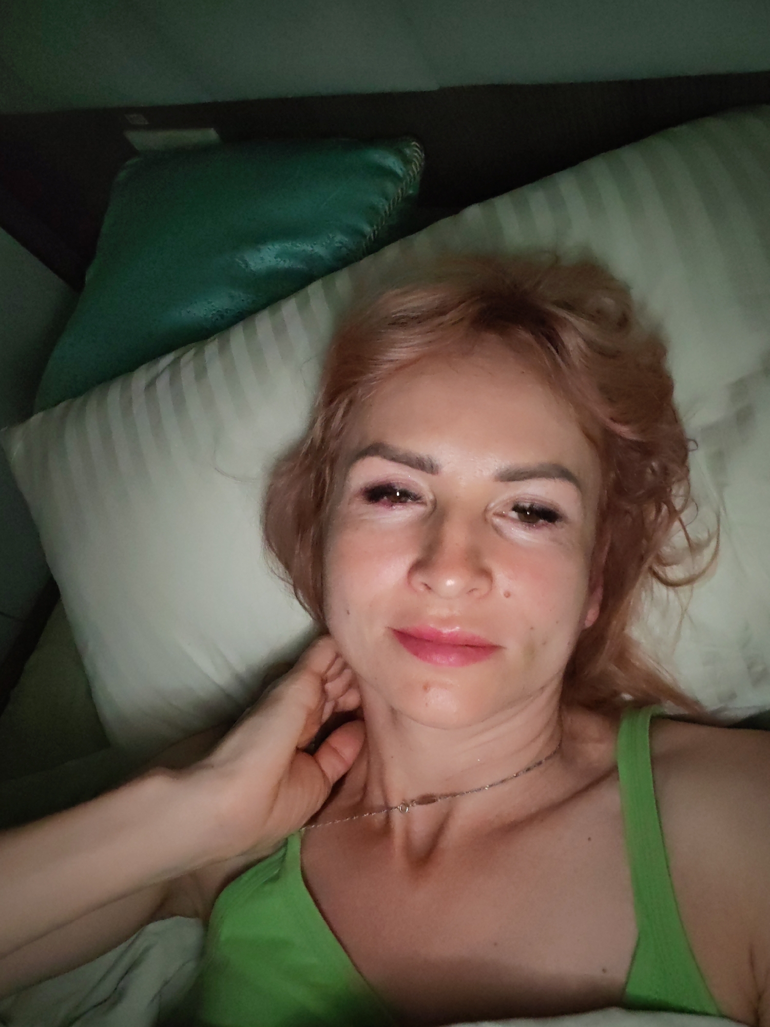 In this close-up, low-light selfie, a woman with light-colored hair and pink lipstick lies on her side in bed, propped up by pillows. She is wearing a green top and a delicate necklace. Her right hand rests on her neck, and her gaze is directed towards the camera. The background consists of a headboard, a dark green decorative pillow, and a beige pillow with white stripes. The scene appears to be indoors, likely in a bedroom. There is no information in the image to suggest a location in Yoshkar-Ola, Russia.