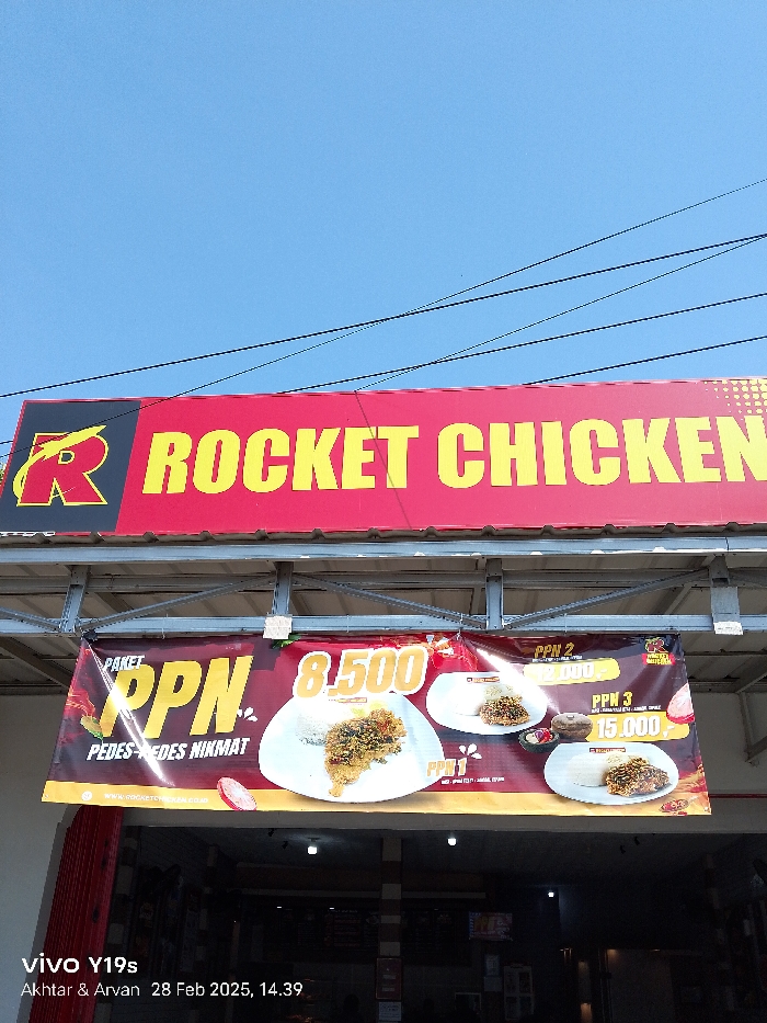 The image shows a storefront for a fried chicken restaurant called "Rocket Chicken". The storefront has a large red and yellow sign above a banner that advertises the "Paket PPN" (PPN Package) with the prices listed. There are several overhead power lines in the sky. The location of this storefront is on Jl. Raya Patrol, Patrol, Indramayu Regency, West Java, Indonesia.