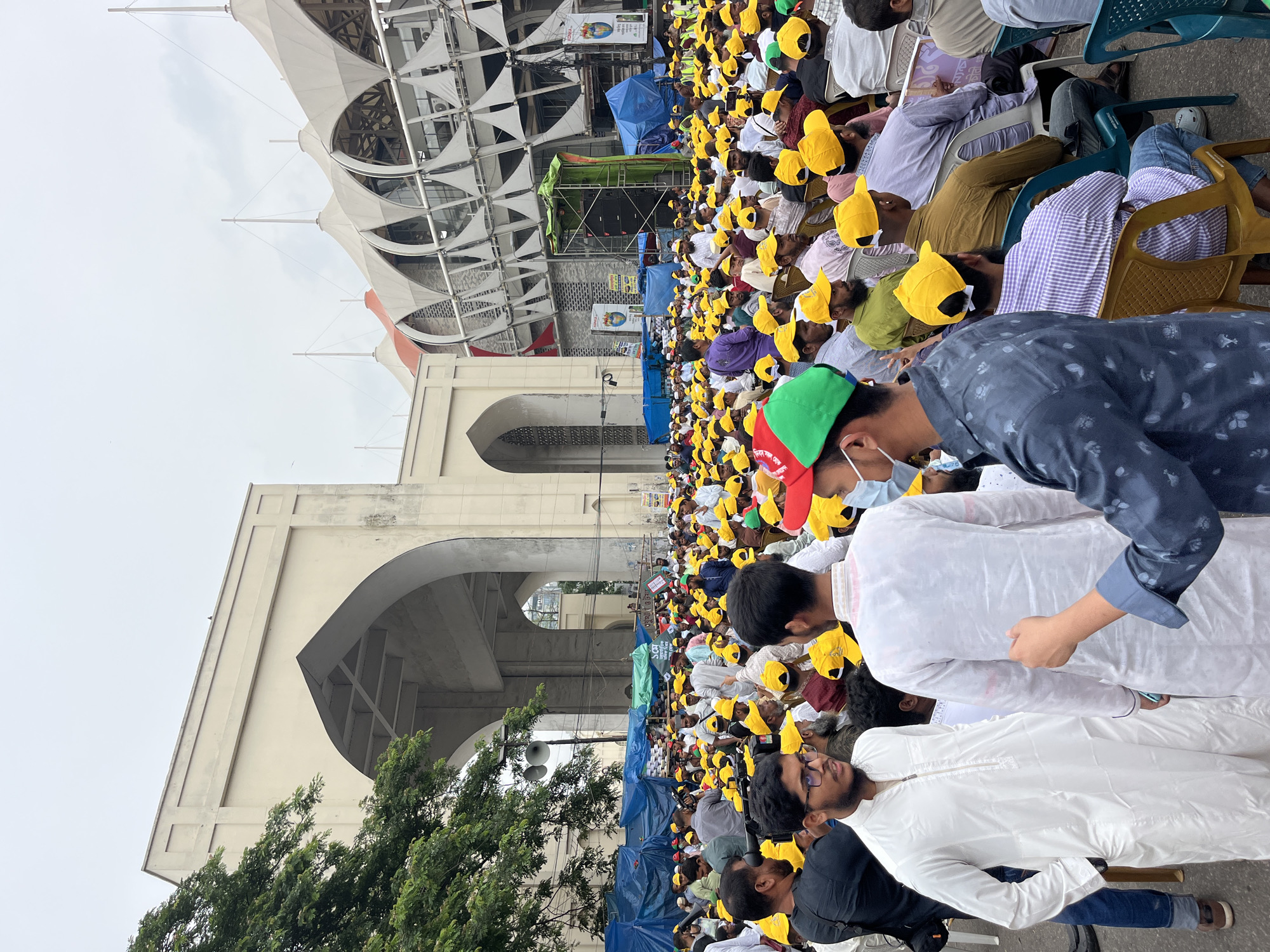 A large crowd, predominantly men, wearing yellow topi (caps) and white clothing are seated on blue plastic chairs during a public gathering in Dhaka, Bangladesh. The Baitul Mukarram National Mosque's architecture, with its large arches and distinctive white structure, is visible in the background. A stage with speakers and blue canopies is set up amidst the crowd. Some individuals are wearing face masks, and some are standing or walking around. Green trees are visible in the foreground. The sky is overcast.