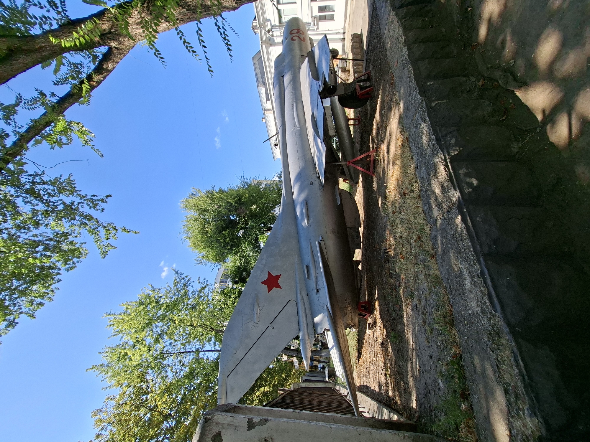 A Soviet-era fighter jet, painted in a light gray, is on display outdoors. The jet has a red star insignia on its tail and the number "29" in red on its nose. It is elevated on red support stands. The jet is situated on a patch of dirt and dry grass, bordered by a low stone wall. In the background, there is a white building with windows and a hint of trees and a clear blue sky. The scene appears to be in a park or museum exhibit area.