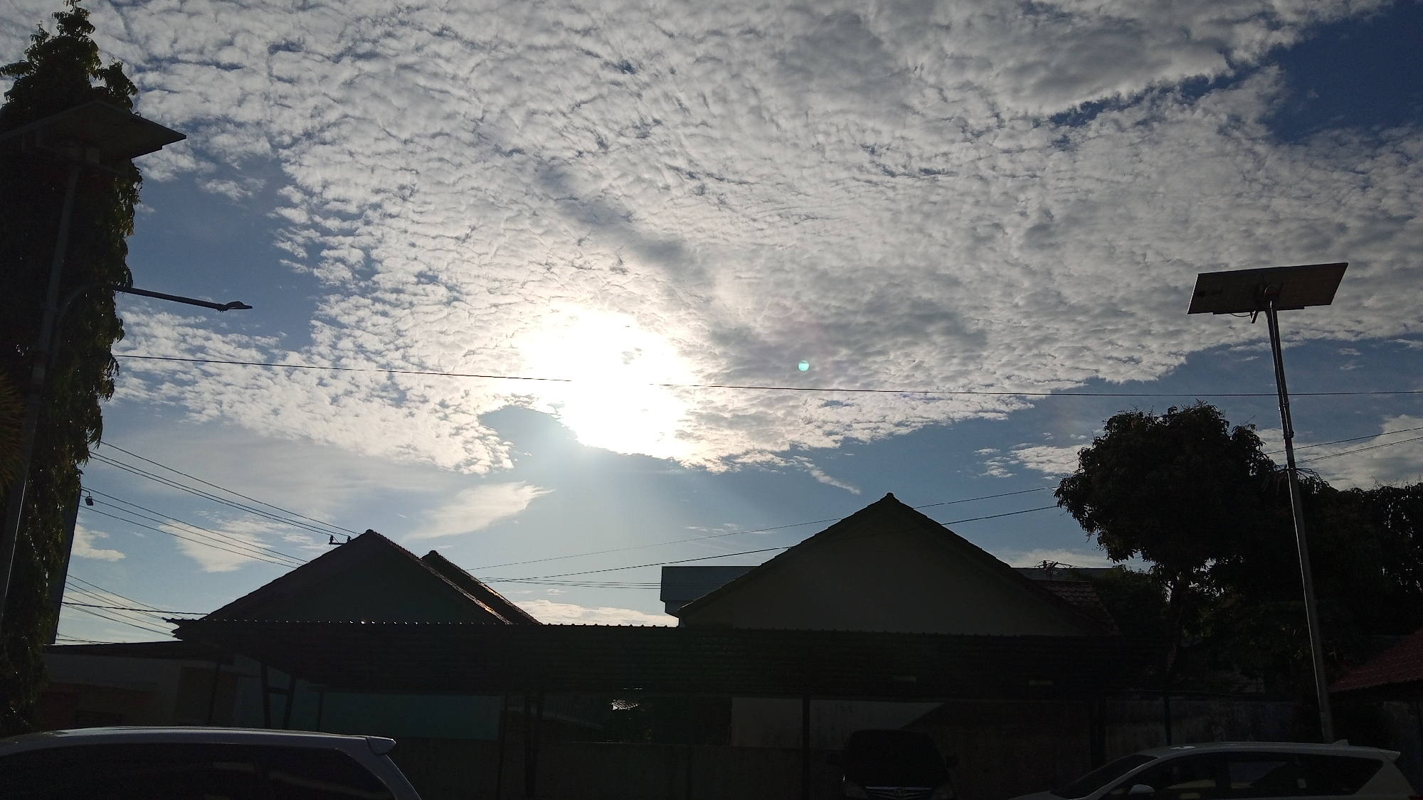 The image depicts a street scene in Bireuen, Indonesia, under a partially cloudy sky. The sun is visible, partially obscured by scattered clouds, casting a bright light. In the foreground, the dark silhouettes of two houses with prominent triangular roofs are visible. Utility wires are strung across the sky, supported by poles. A tall, slender street light with a solar panel at its apex stands on the right side of the frame. To the left, a tall, leafy tree is silhouetted against the sky. The lower portion of the image shows the top of a white vehicle, also in silhouette. The overall impression is that of a residential area during daylight hours, possibly early morning or late afternoon, given the lighting conditions.