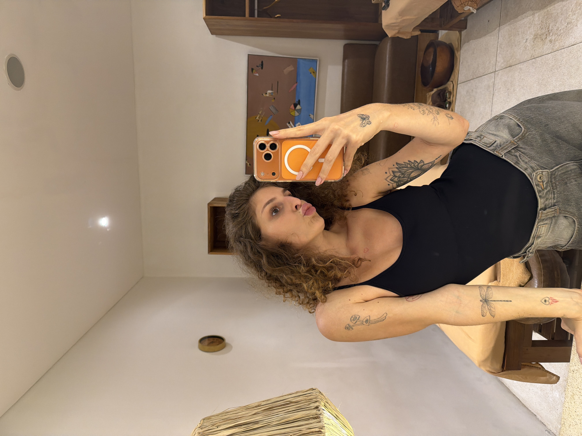 A young woman with curly brown hair, wearing a black tank top and grey jeans, stands in a room and takes a selfie in a mirror. She is pouting her lips and holding an orange-cased phone in her right hand. Tattoos are visible on her arms and collarbone, including a dragonfly on her left forearm and a butterfly on her right wrist. The room appears to be a spa or wellness center, with a massage table draped in a tan cloth in the background. A wooden cabinet with shelves is to the right, and a decorative lamp with a woven shade is to the left. A painting depicting figures on a beach hangs on the wall behind her. The flooring is tiled. The location is identified as North Kuta, Indonesia.
