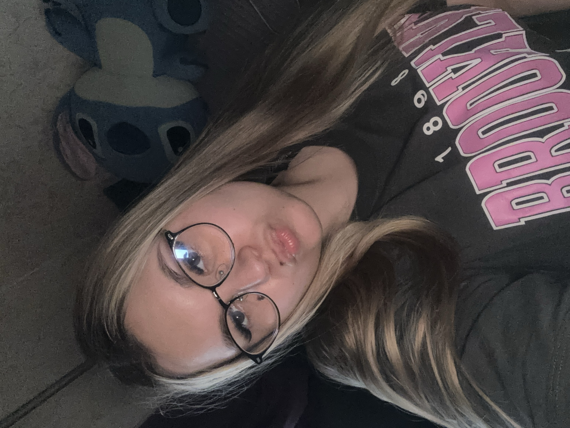 A young woman with blonde hair and glasses is taking a selfie. She is wearing a dark t-shirt with the words "BROOKLYN 1898" printed on it in pink lettering. Behind her, a blue plush toy resembling Stitch from the Disney movie "Lilo & Stitch" is visible. The lighting is dim, suggesting an indoor setting. There is no information available about the specific location in the city of Bolshoi Bukor, Russia.
