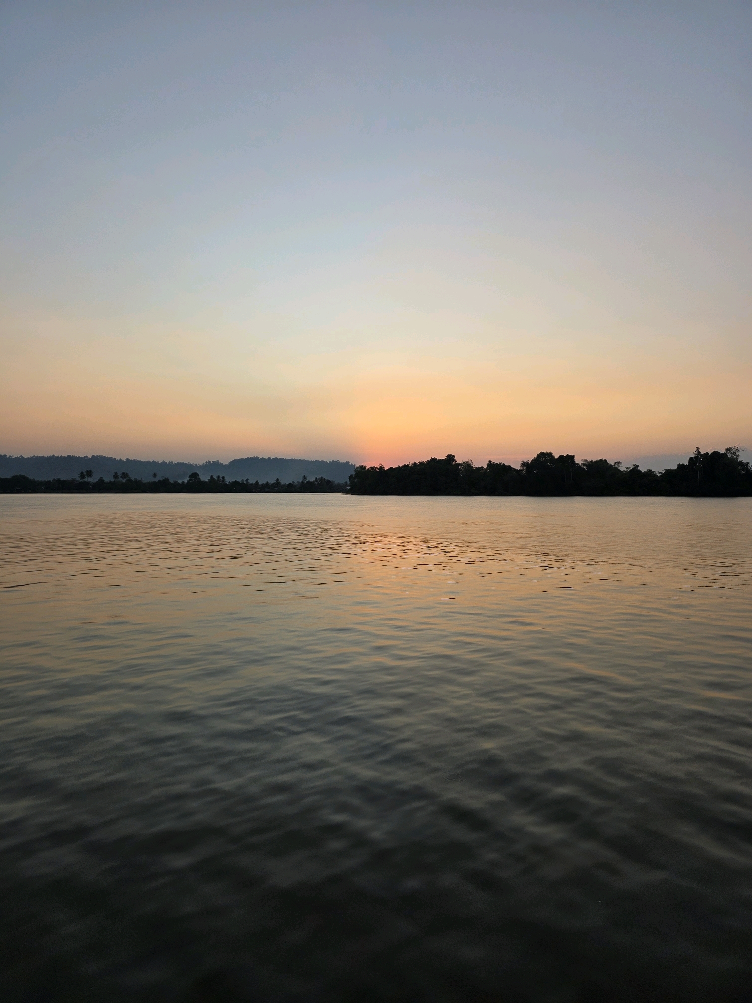 A wide shot captures a tranquil river scene at sunset in Kota Tanjung Selor, Indonesia. The sky displays a gradient of soft blues and oranges, with the sun just dipping below the horizon, casting a warm glow. A dense, dark treeline stretches across the middle ground, silhouetted against the vibrant sky. The foreground is dominated by the calm river, its surface rippled by gentle waves that reflect the sunset hues. The overall atmosphere is peaceful and serene.