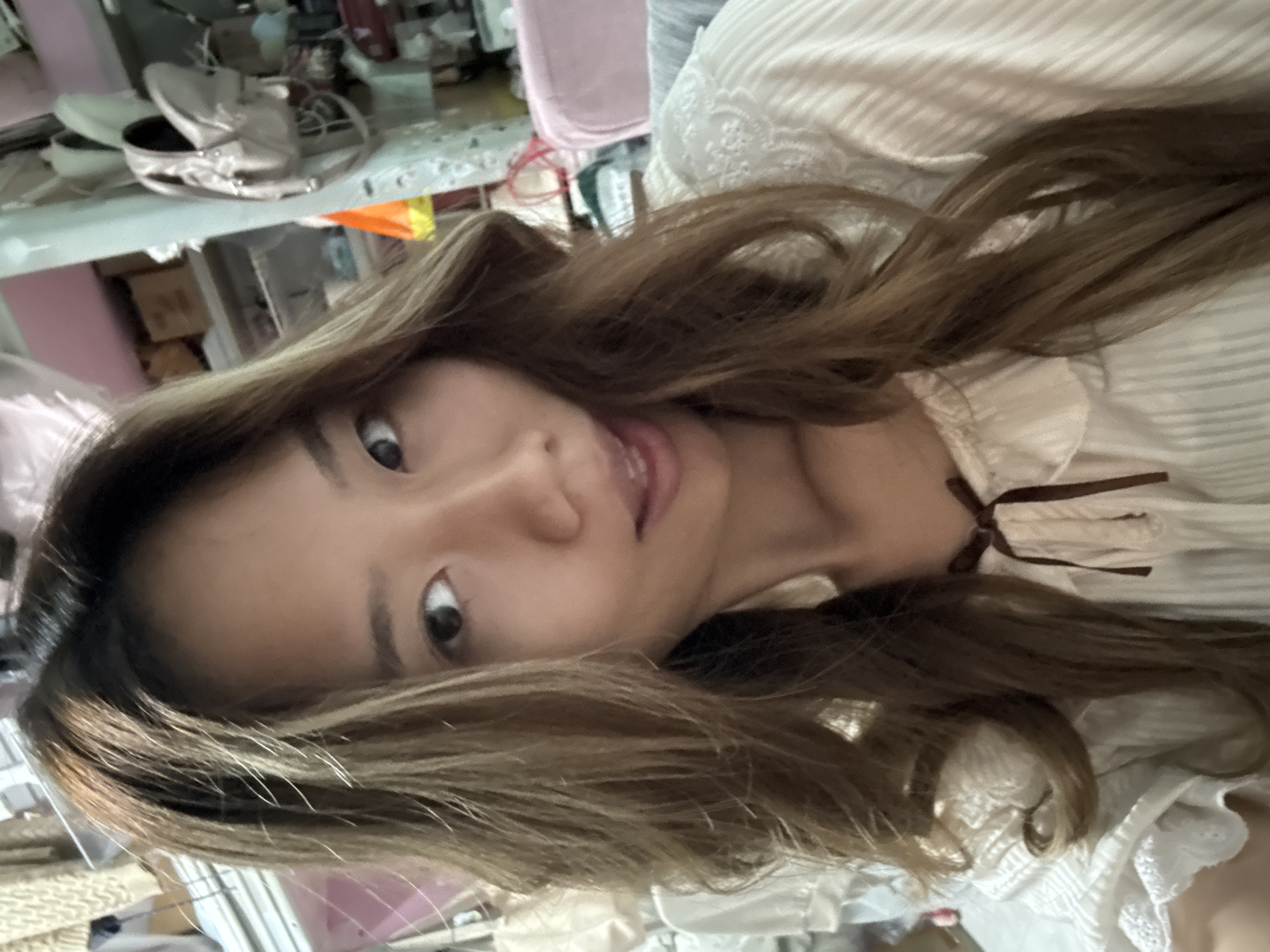 A woman with long brown hair looks at the camera. She is wearing a white top with a brown ribbon at the neck. The background is cluttered, with items on a shelf and a pink object. It's difficult to determine the exact location in Nan'an District, China, without more information or context.