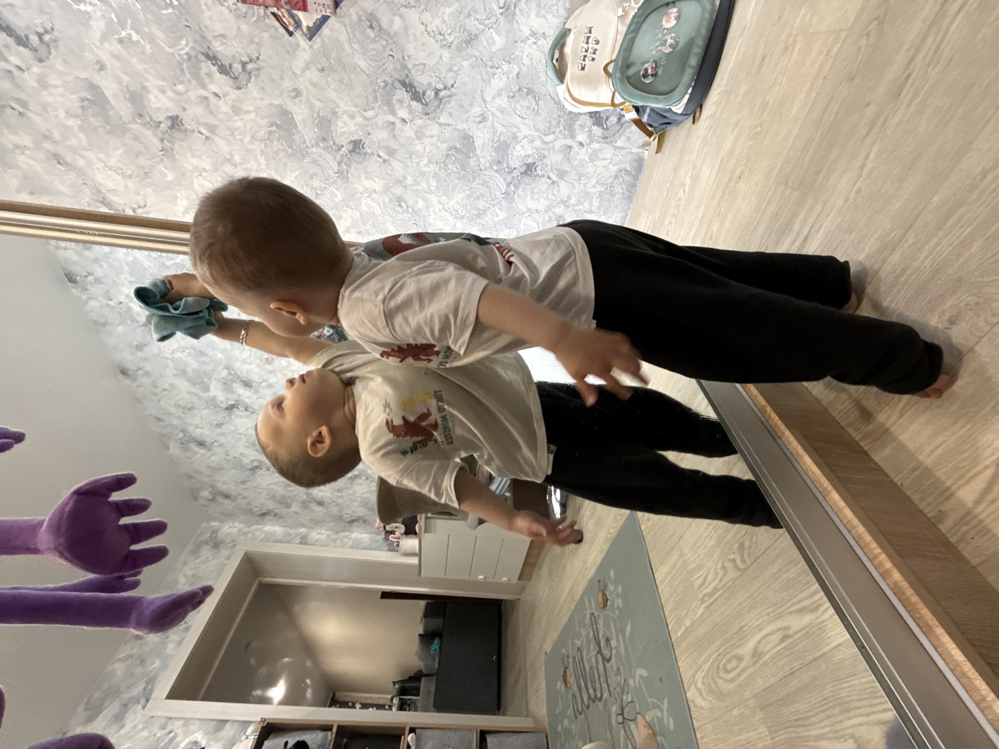 A child is cleaning a mirror with a blue cloth. The room appears to be a bedroom with a wooden floor, patterned wallpaper, and a purple stuffed animal. Other visible objects include a backpack, a rug, a dresser, and storage units. The location may be Rusanovo, Russia, but this cannot be determined with certainty from the image alone.