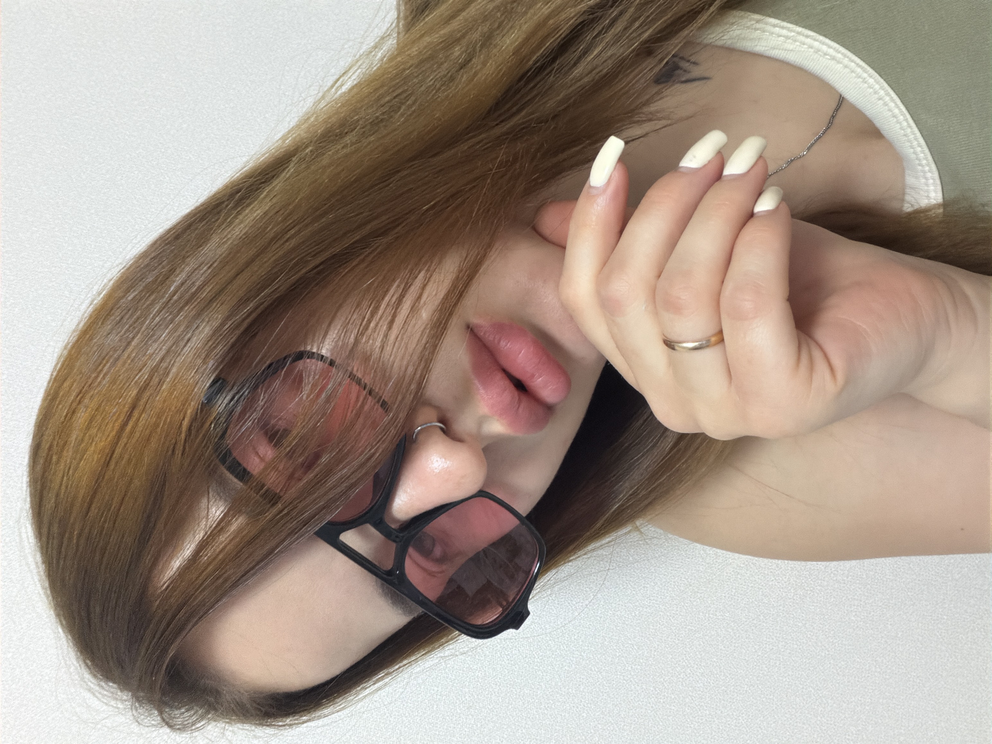 A close-up, high-angle shot captures a person, likely female, with long, flowing light brown hair. Their head is tilted, and their right hand is raised, partially obscuring the left side of their face, specifically the area around their eye and temple. The nails on the visible hand are long, square-shaped, and appear to be painted in a light, possibly white or pale yellow, color. A gold-colored ring adorns the ring finger of this hand.
The person is wearing small, square-framed sunglasses with dark frames and reddish-tinted lenses, positioned on their nose. A small silver hoop nose ring is visible on their right nostril. Their lips are slightly parted, showcasing a full shape and a pinkish hue from lipstick or gloss. A thin silver chain necklace is draped around their neck, partially covered by their hair and a light green garment with a ribbed neckline. Faintly visible beneath the hair on the upper shoulder or neck area is a dark, abstract-looking tattoo.
The background is a plain, textured light-colored surface, suggesting an indoor setting. No specific activity or event is taking place beyond posing for the photograph. The time of day and weather are not discernible, and there are no explicit location cues. The overall mood is stylish and somewhat enigmatic due to the obscured face. No text is visible in the image.
