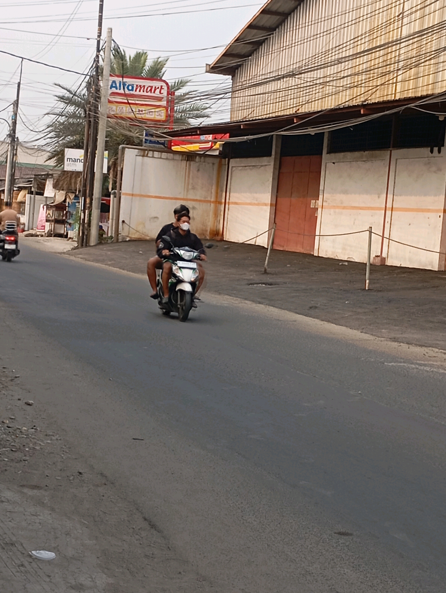 In Babelan, Indonesia, a medium shot captures two individuals riding a white and green scooter along a paved road. The rider in front wears a black shirt and a white face mask, while the passenger behind also wears a black shirt and a dark head covering. To the left of the road, a sign with the "Alfamart" logo is visible, situated on a utility pole amidst a tangle of electrical wires and a palm tree. Further back on the left, a person is seen on another scooter. The right side of the image shows the side of a building with a corrugated metal roof and a red door, bordered by a white wall with orange stripes. A rough, dirt shoulder runs alongside the road in front of the building, marked by two wooden posts connected by a rope. The road surface is asphalt with some debris scattered on it. The sky is overcast.