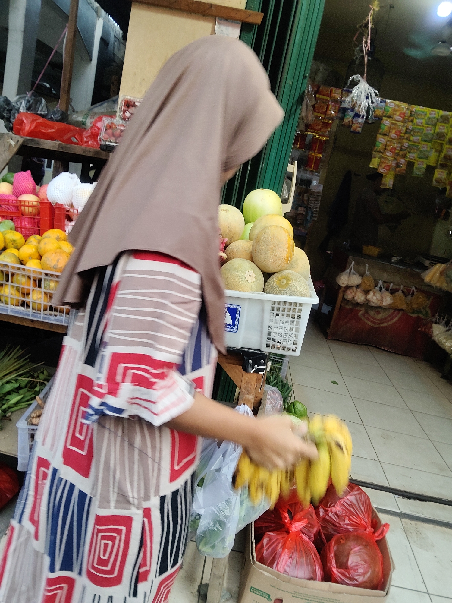 A medium shot captures a woman wearing a hijab and a patterned dress in what appears to be a market stall in Mekarmukti, Indonesia. She is holding a bunch of bananas with her right hand, and her left arm is extended towards a box filled with red plastic bags containing round objects. The market stall is stocked with various fruits, including oranges in a white basket, and melons in another white basket. Behind the stall, shelves display numerous packets of food items. The ground is tiled, and a green metal gate is visible to the left. The lighting suggests it is daytime.