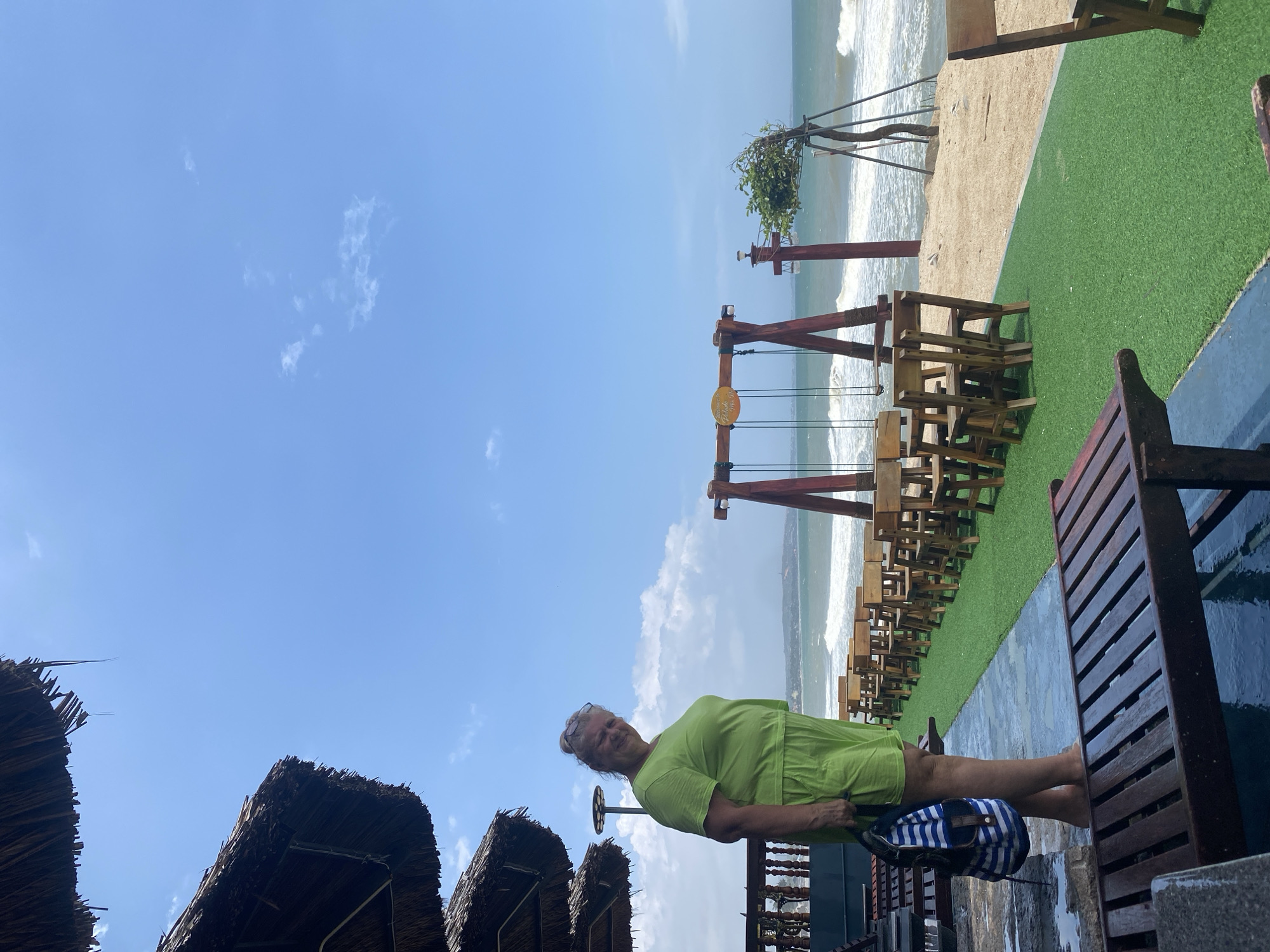 A woman in a bright green t-shirt and shorts stands on a wet stone patio, looking at the camera with a smile. She holds a blue and white striped bag in her left hand. Behind her, a row of empty wooden chairs and tables sits on a patch of artificial turf, leading to a sandy beach where waves crash. A wooden swing set with a sign hangs on the beach. In the background, the ocean stretches to the horizon under a bright blue sky with scattered clouds. Thatch-roofed structures are visible in the upper left corner of the frame. The scene appears to be at a beachfront establishment in Mũi Né, Vietnam.