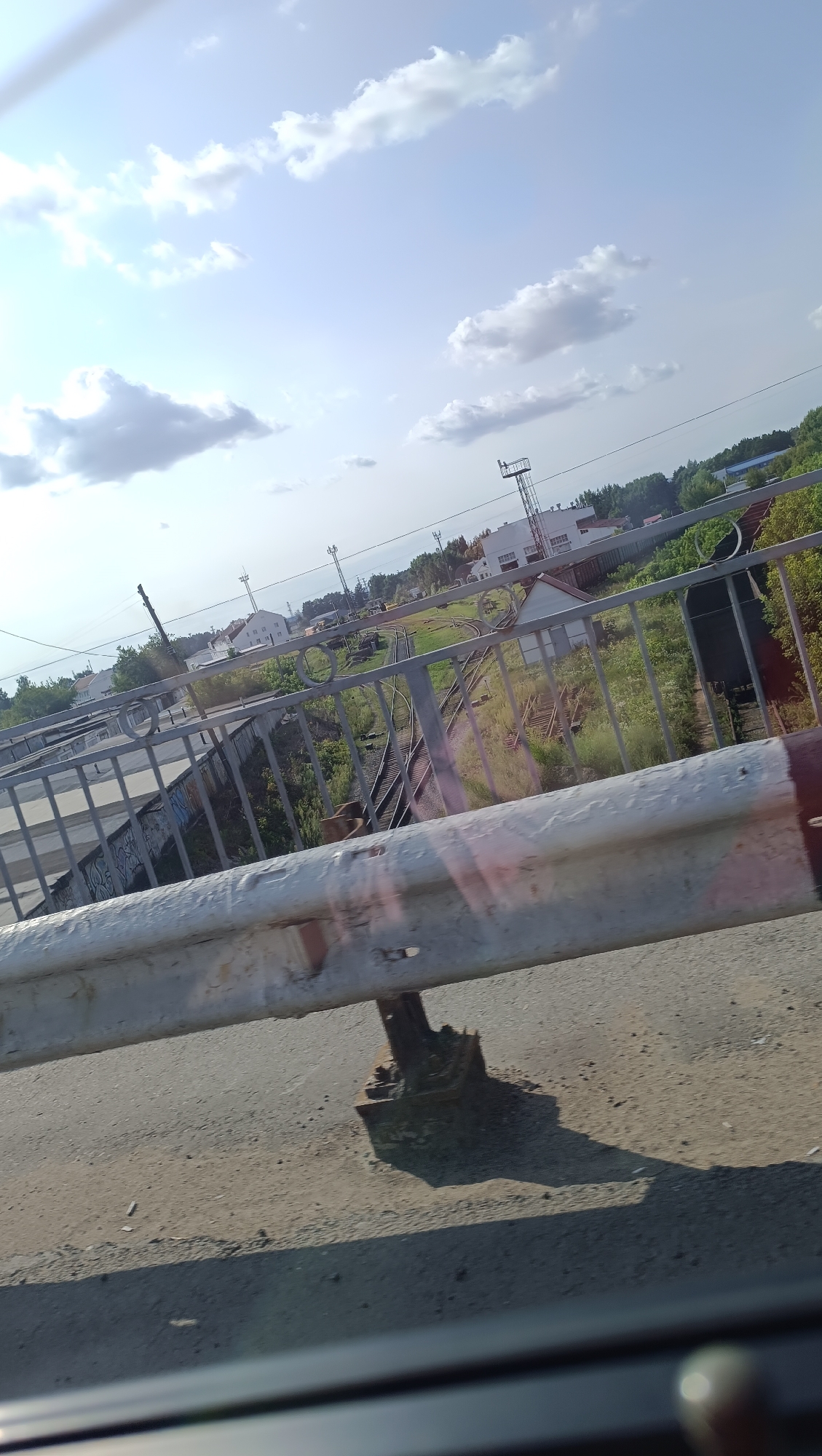 The image captures an outdoor scene from what appears to be a moving vehicle, looking towards a sky with scattered clouds. In the foreground, a weathered, white metal guardrail runs horizontally across the bottom of the frame, supported by a rusty metal post with a complex base structure. The road surface beneath the guardrail is asphalt, with some debris scattered on it. Beyond the guardrail, a metal fence with vertical bars and circular decorative elements borders an area with railway tracks. Various industrial buildings, some with white facades, are visible in the mid-ground, interspersed with trees and greenery. A tall, lattice-like structure, possibly a pylon or radio tower, stands to the right of the frame, and another similar structure is visible further in the distance. The overall environment suggests an industrial or semi-industrial area within a city. The perspective and angle suggest the photograph was taken from a slightly elevated position, such as from a bridge or overpass, looking down and across the landscape. The lighting indicates it is daytime, with the sun casting shadows. The presence of railway infrastructure suggests this area could be near a train station or industrial complex. Given the query about the city of Chaikovsky, Russia, this scene likely depicts a part of its urban or industrial landscape.
